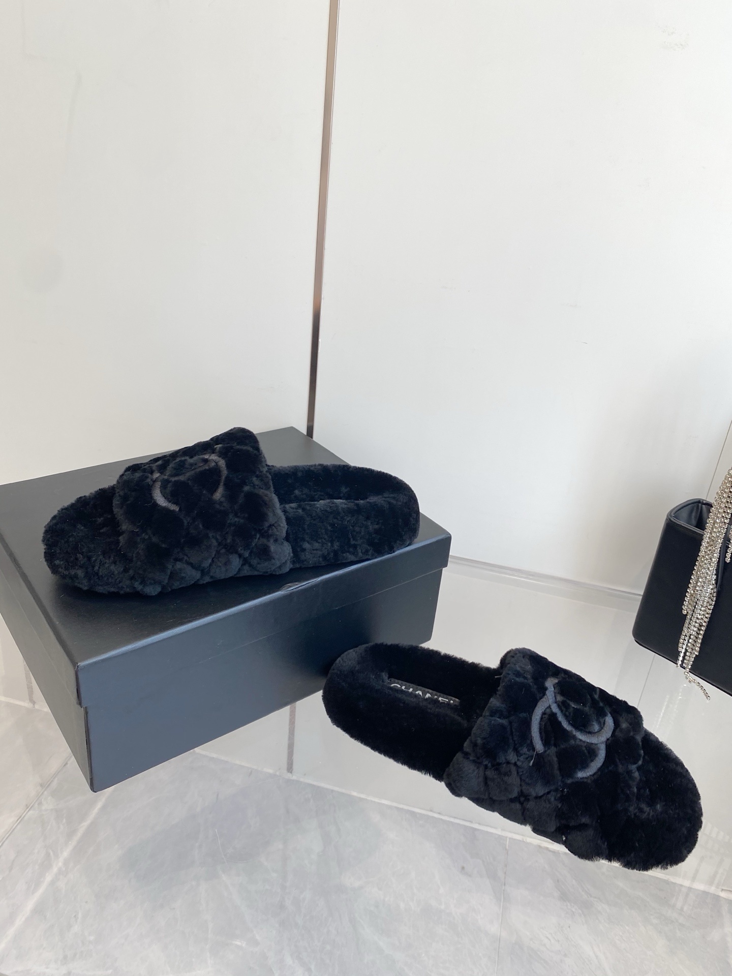 Pair of black plush faux fur slippers with embroidered interlocking C logo on a marble surface near a black handbag. - Hình 7
