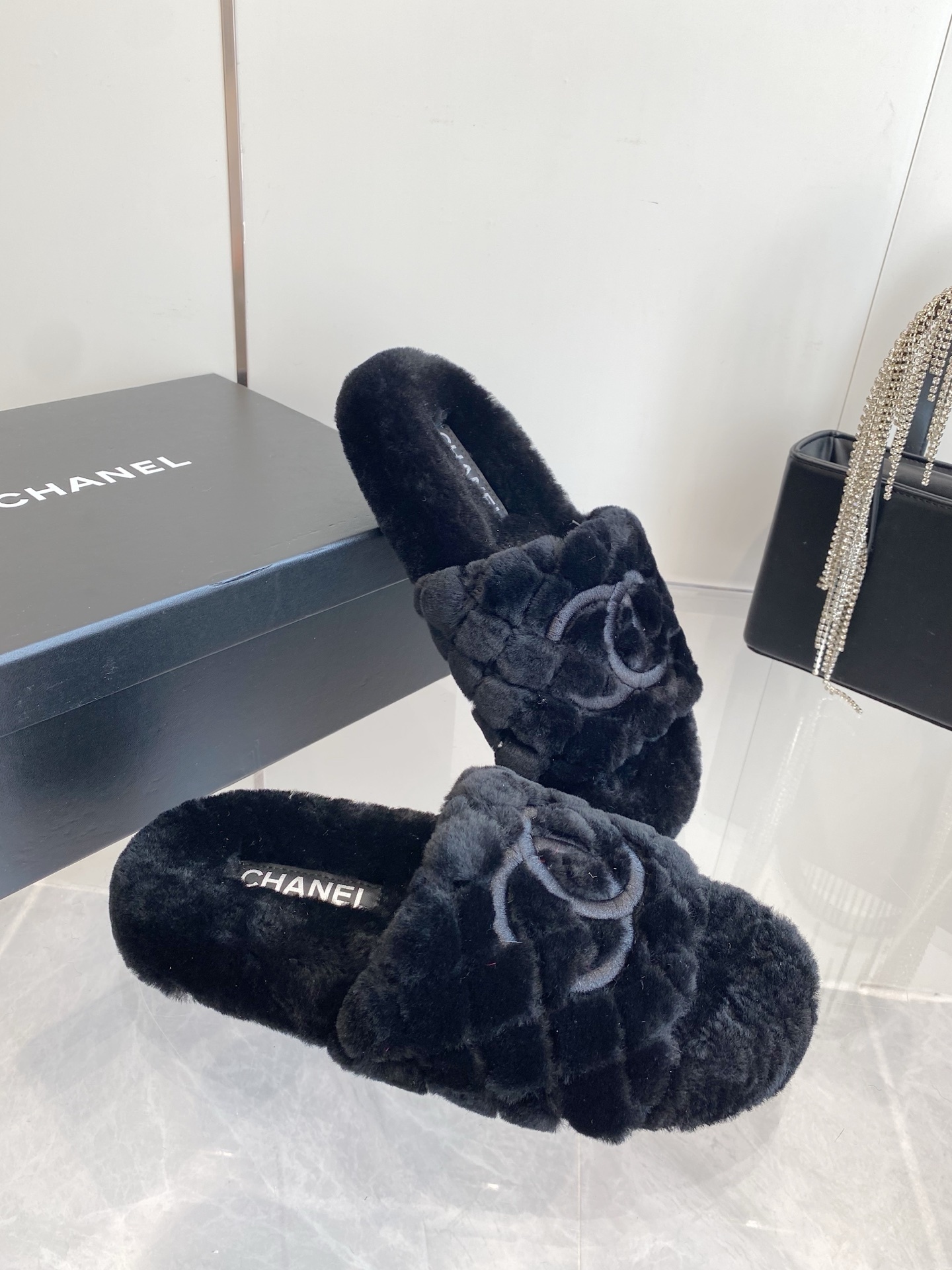 Pair of black plush faux fur slippers with embroidered interlocking C logo on a marble surface near a black handbag. - Hình 6