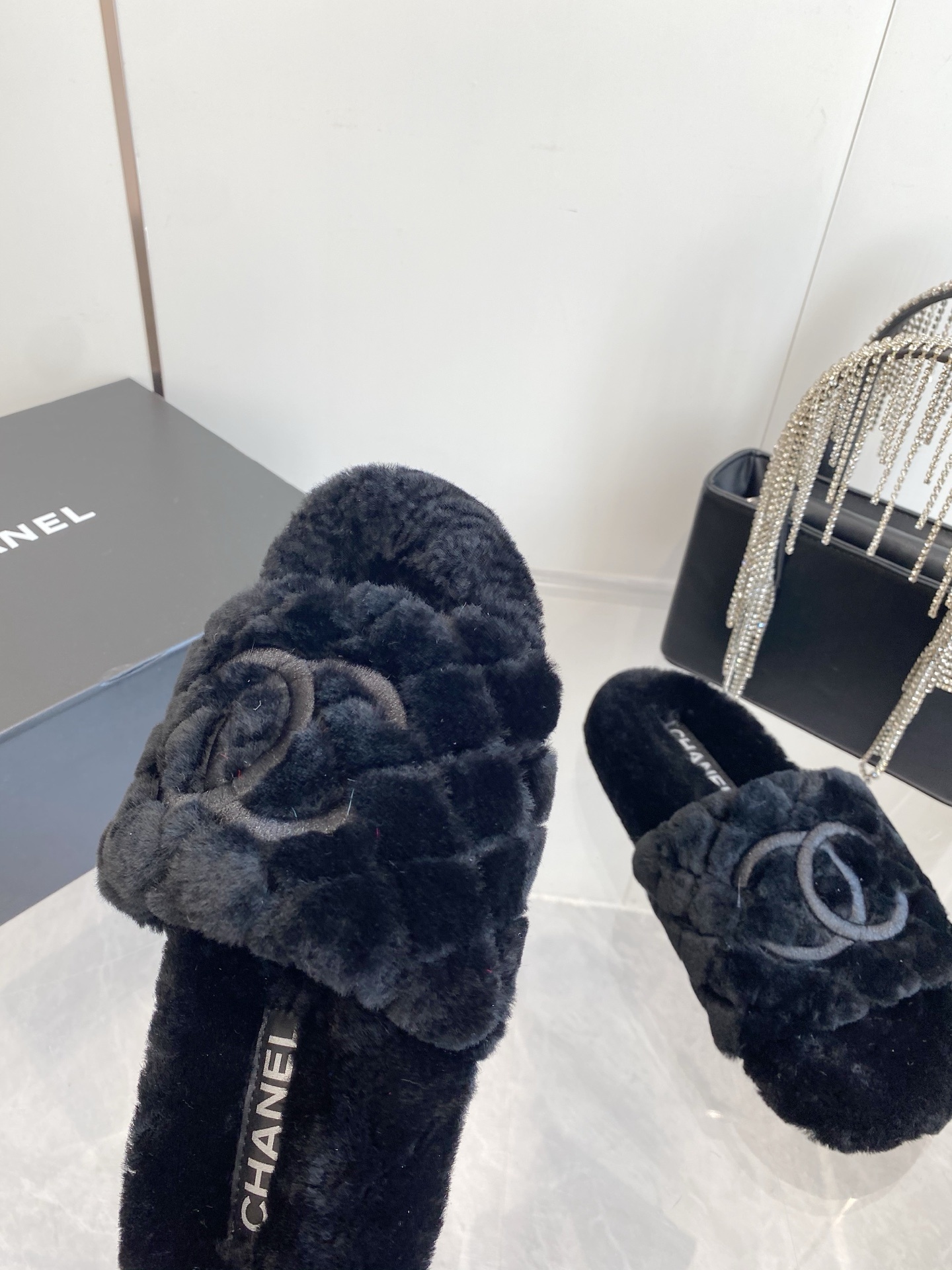 Pair of black plush faux fur slippers with embroidered interlocking C logo on a marble surface near a black handbag. - Hình 5