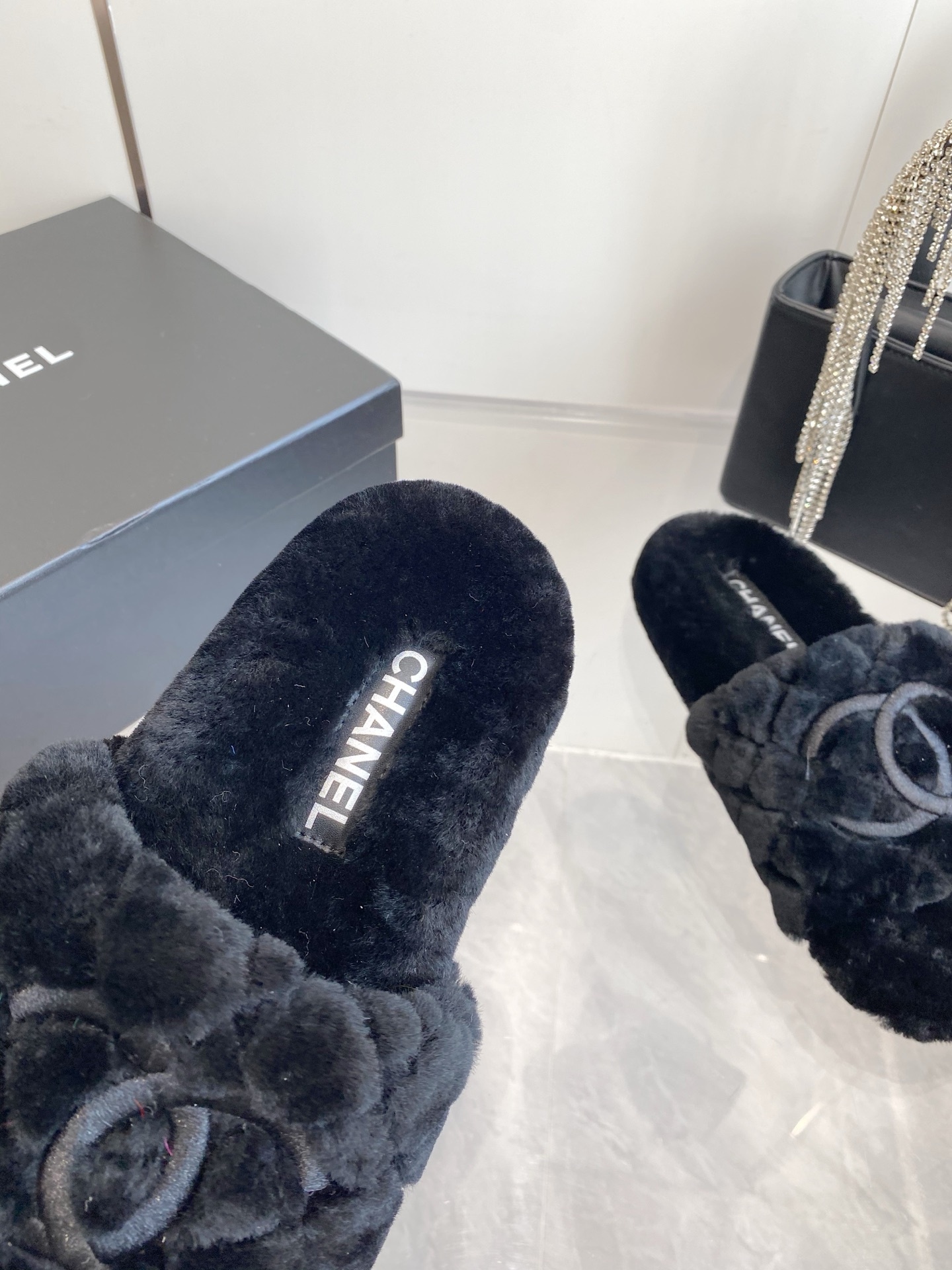 Pair of black plush faux fur slippers with embroidered interlocking C logo on a marble surface near a black handbag. - Hình 4