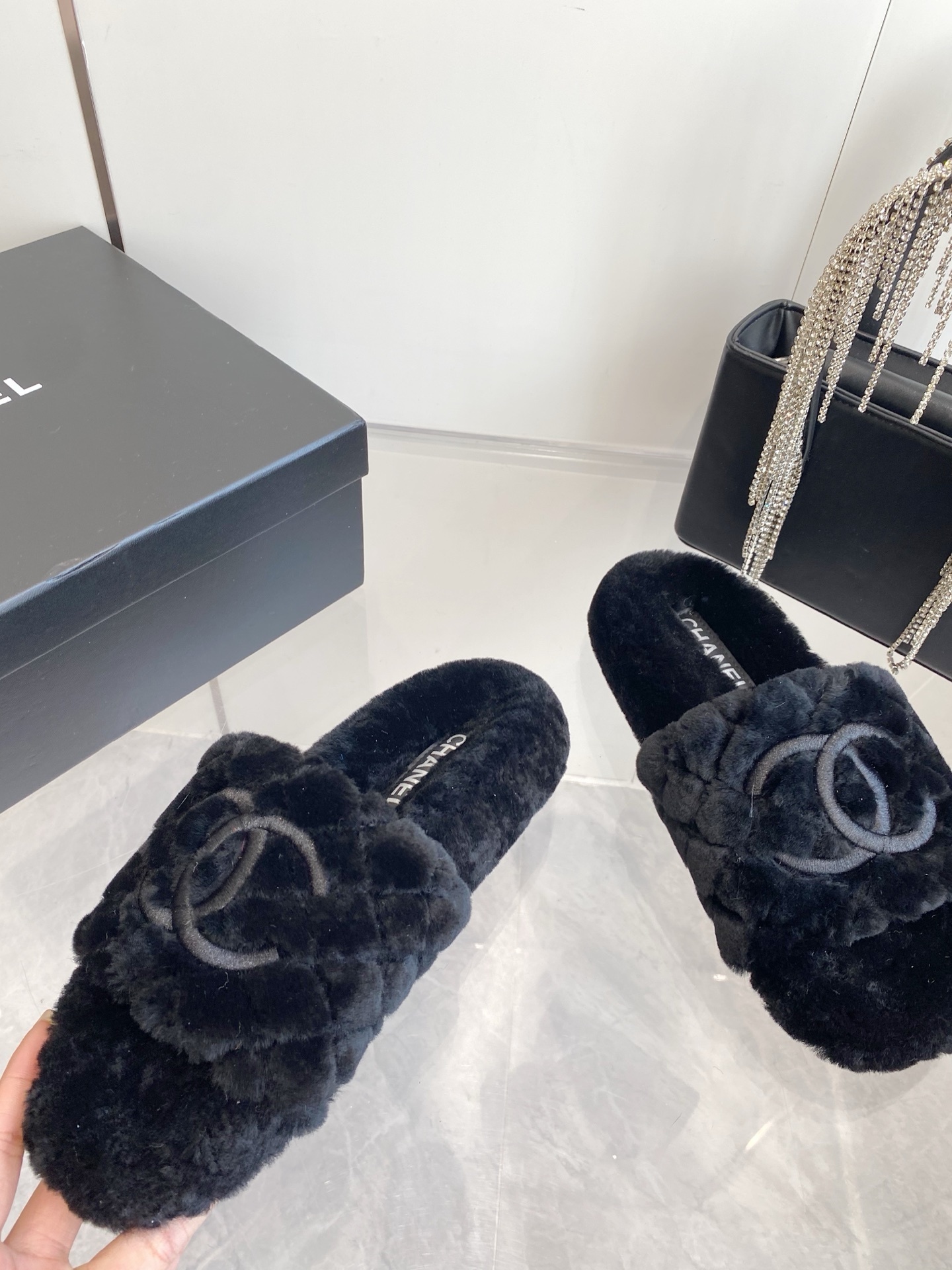 Pair of black plush faux fur slippers with embroidered interlocking C logo on a marble surface near a black handbag. - Hình 3
