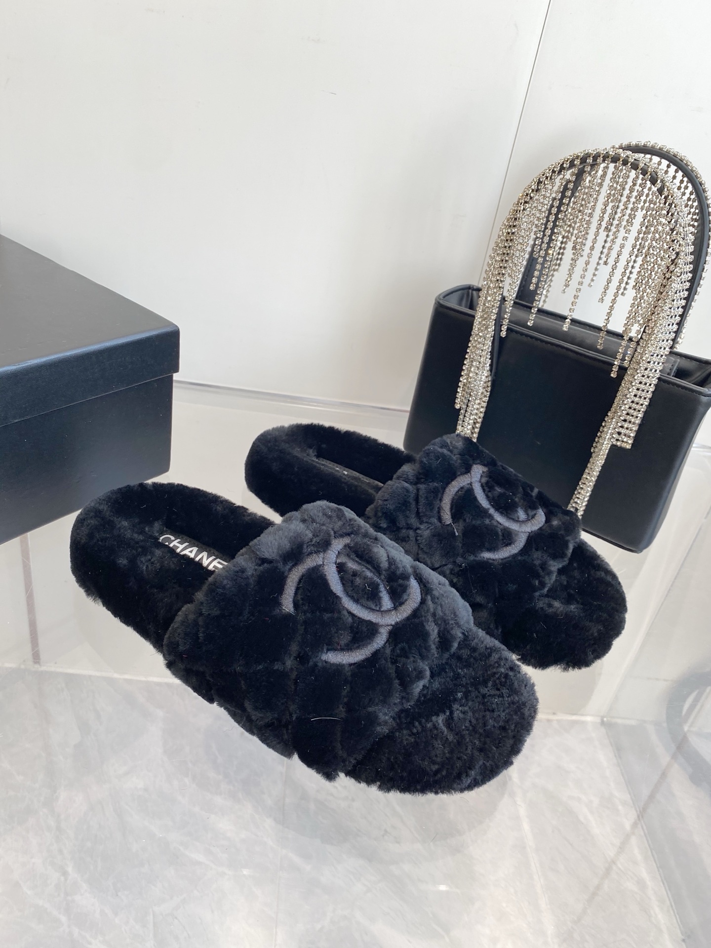 Pair of black plush faux fur slippers with embroidered interlocking C logo on a marble surface near a black handbag. - Hình 2