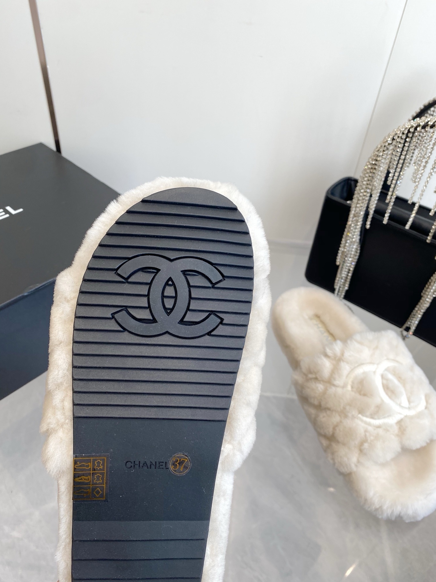 A pair of cream-colored luxury quilted faux fur slippers with an embroidered logo, placed next to a black handbag with crystal fringe. - Hình 9