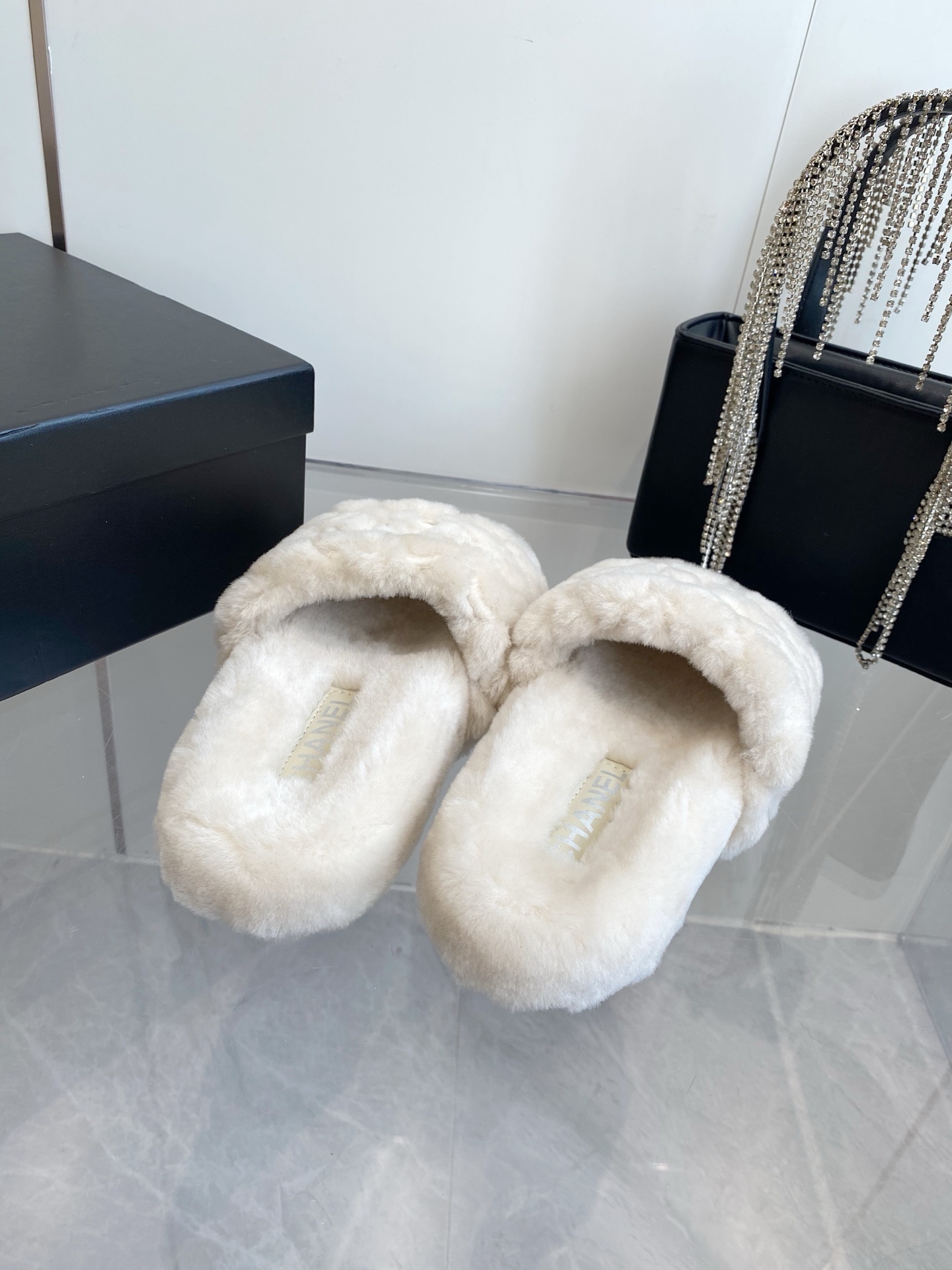 A pair of cream-colored luxury quilted faux fur slippers with an embroidered logo, placed next to a black handbag with crystal fringe. - Hình 8