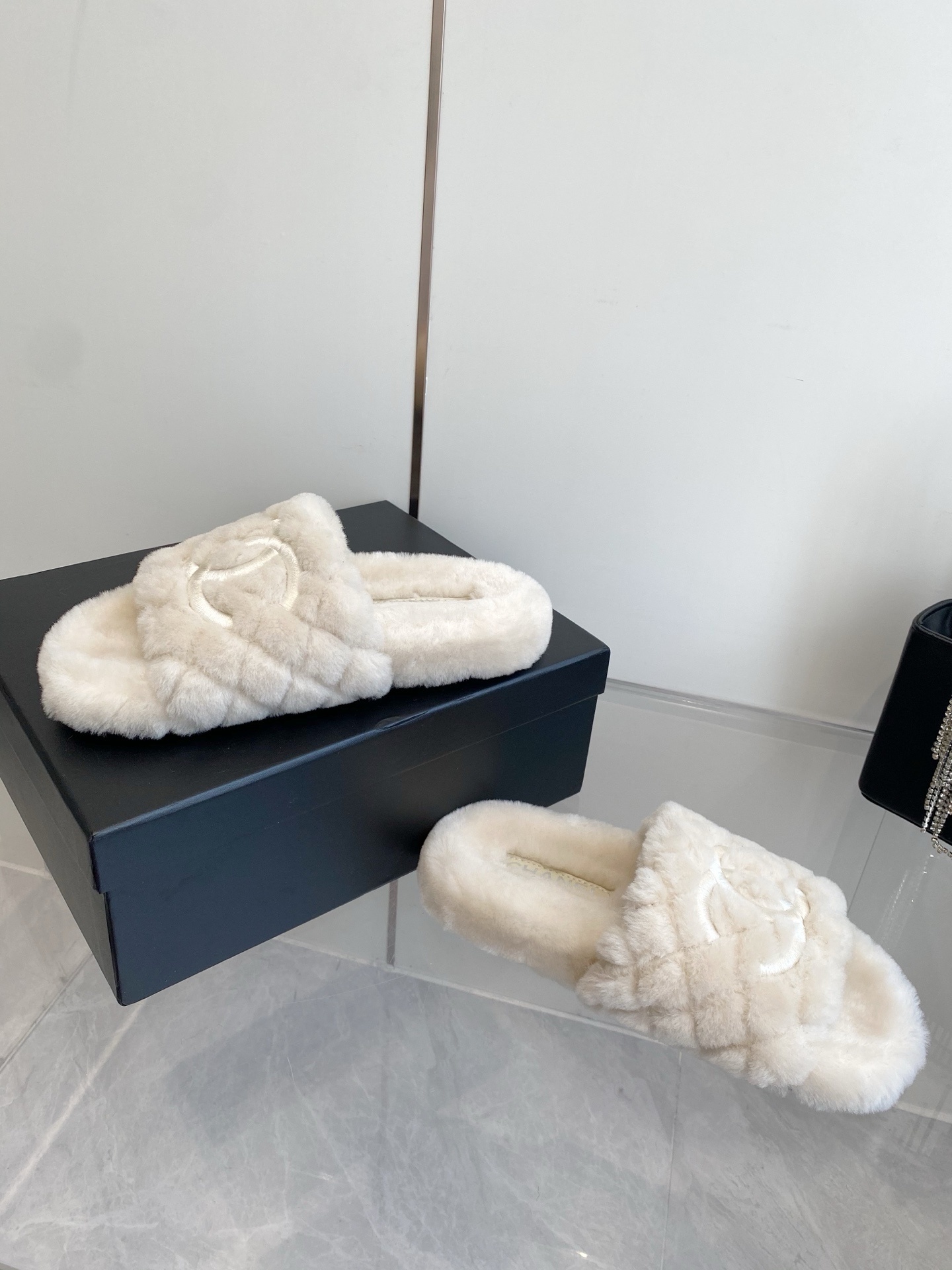 A pair of cream-colored luxury quilted faux fur slippers with an embroidered logo, placed next to a black handbag with crystal fringe. - Hình 7