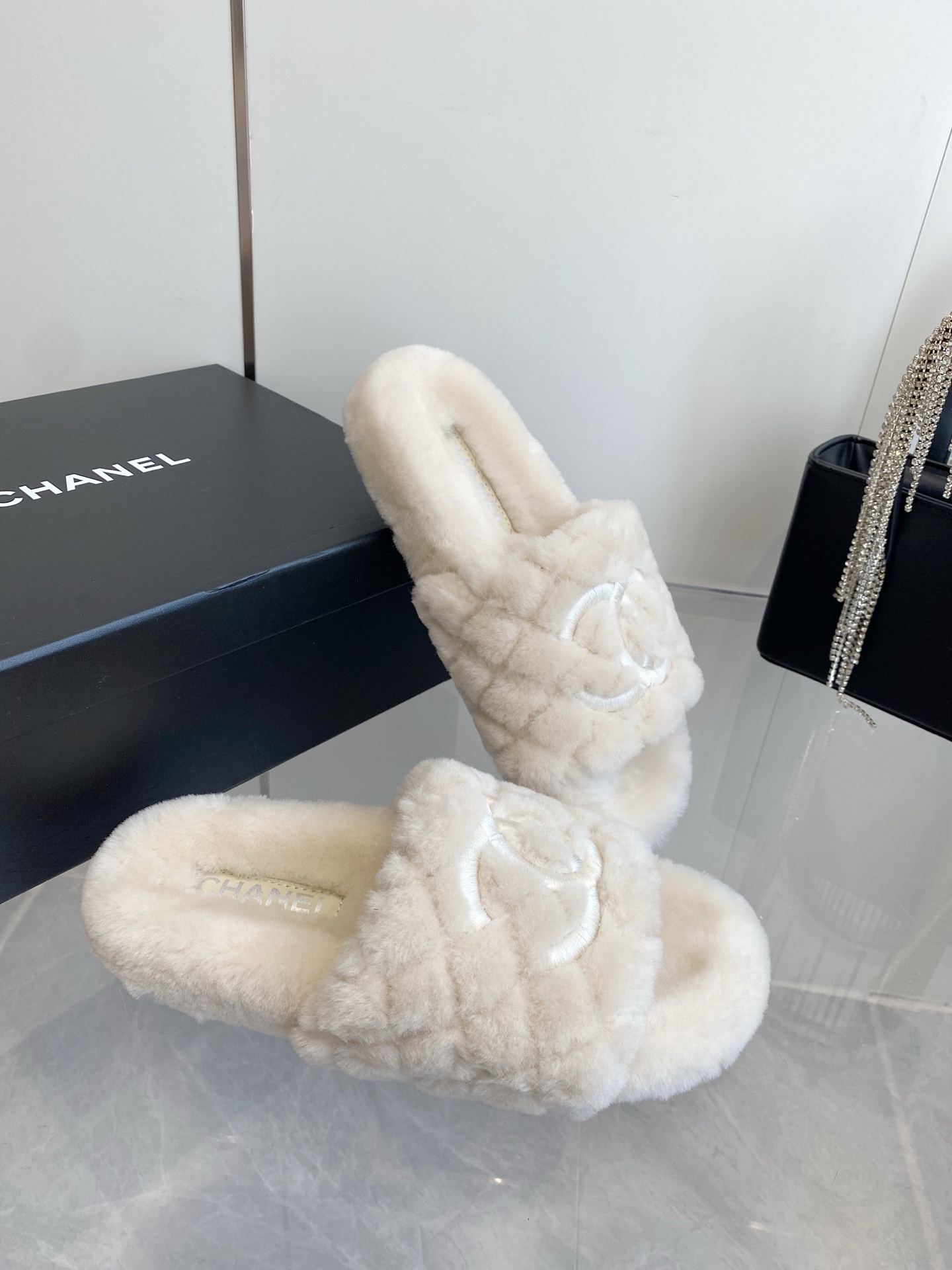 A pair of cream-colored luxury quilted faux fur slippers with an embroidered logo, placed next to a black handbag with crystal fringe. - Hình 6