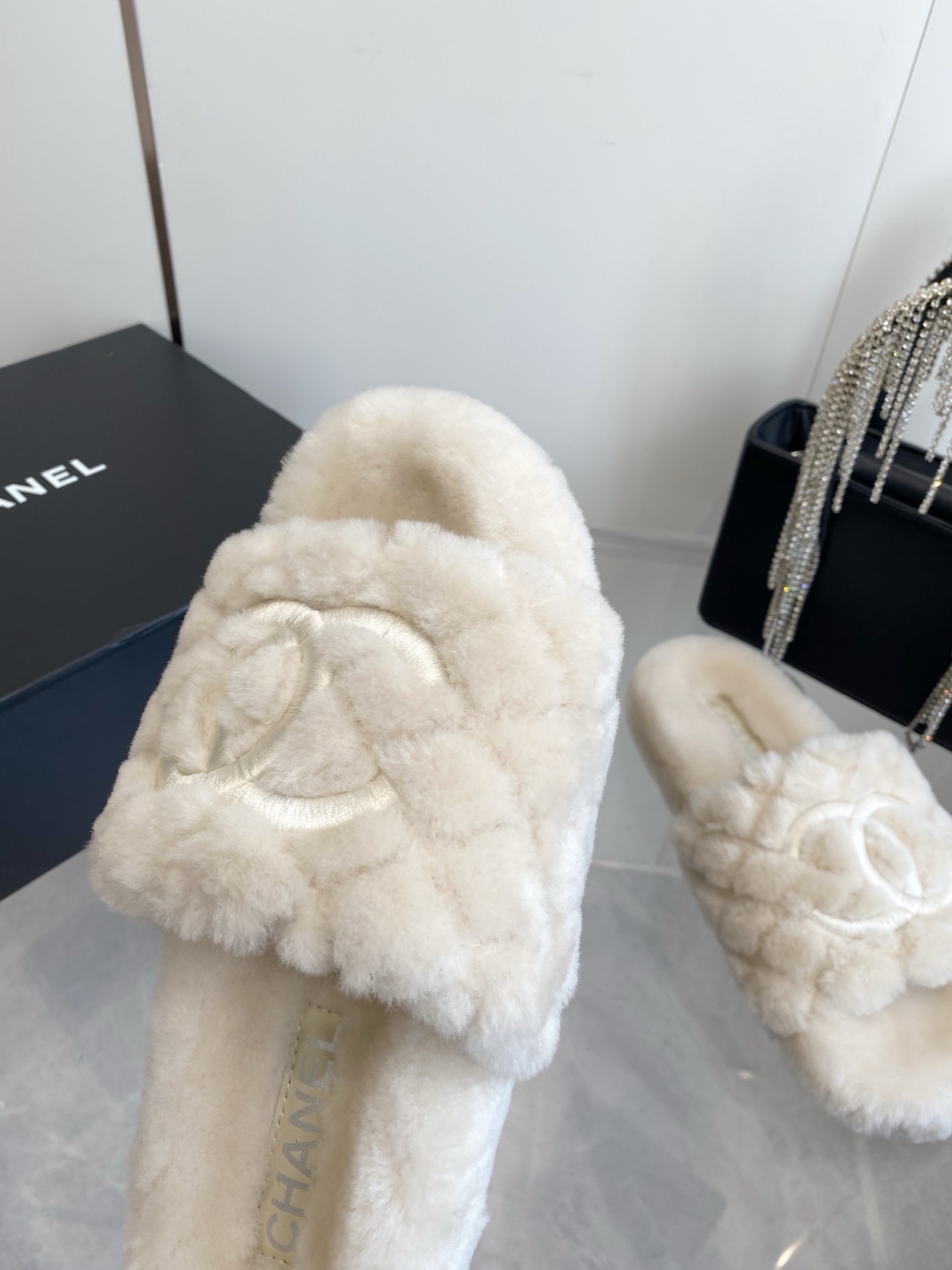 A pair of cream-colored luxury quilted faux fur slippers with an embroidered logo, placed next to a black handbag with crystal fringe. - Hình 5