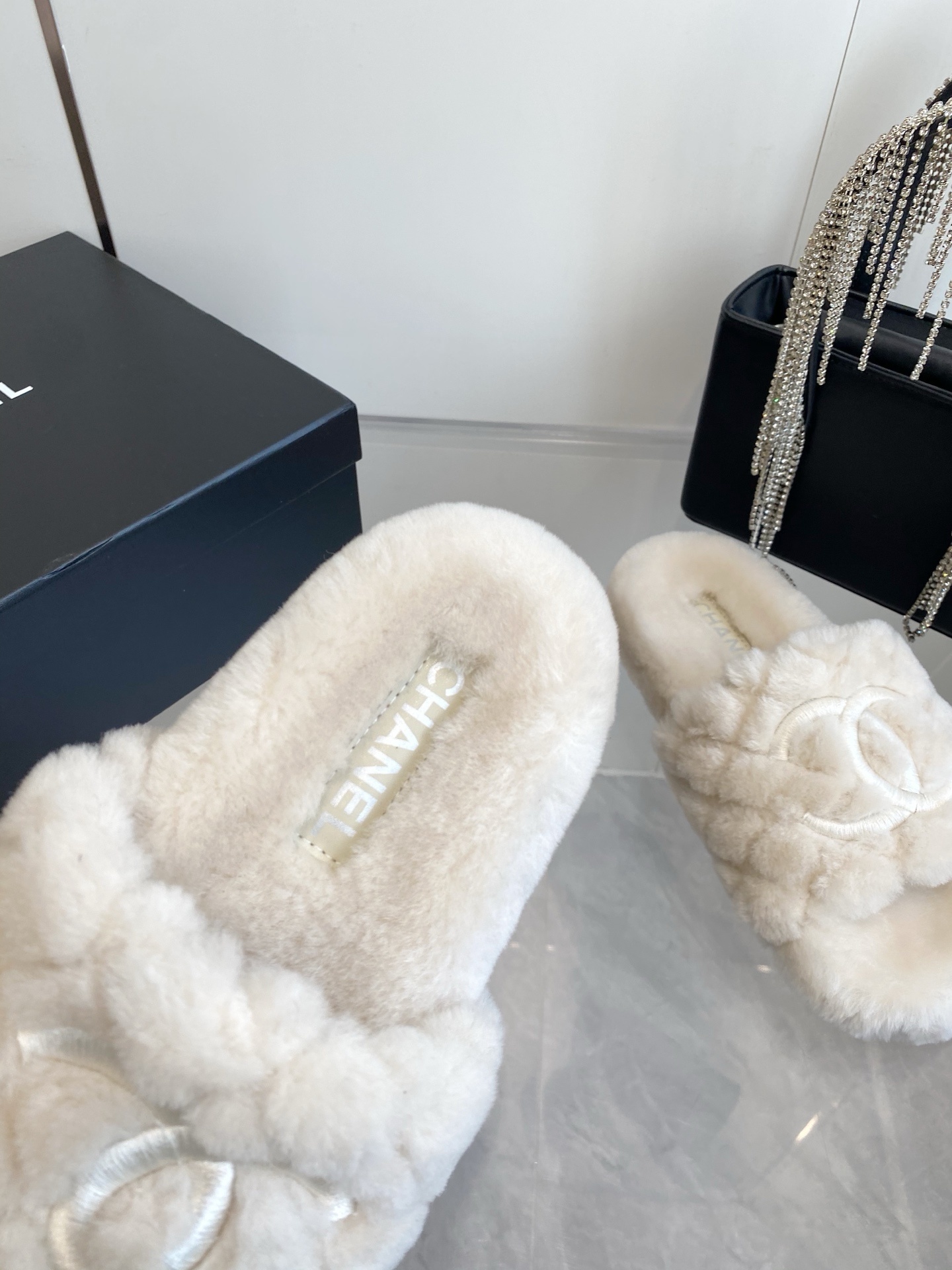 A pair of cream-colored luxury quilted faux fur slippers with an embroidered logo, placed next to a black handbag with crystal fringe. - Hình 4