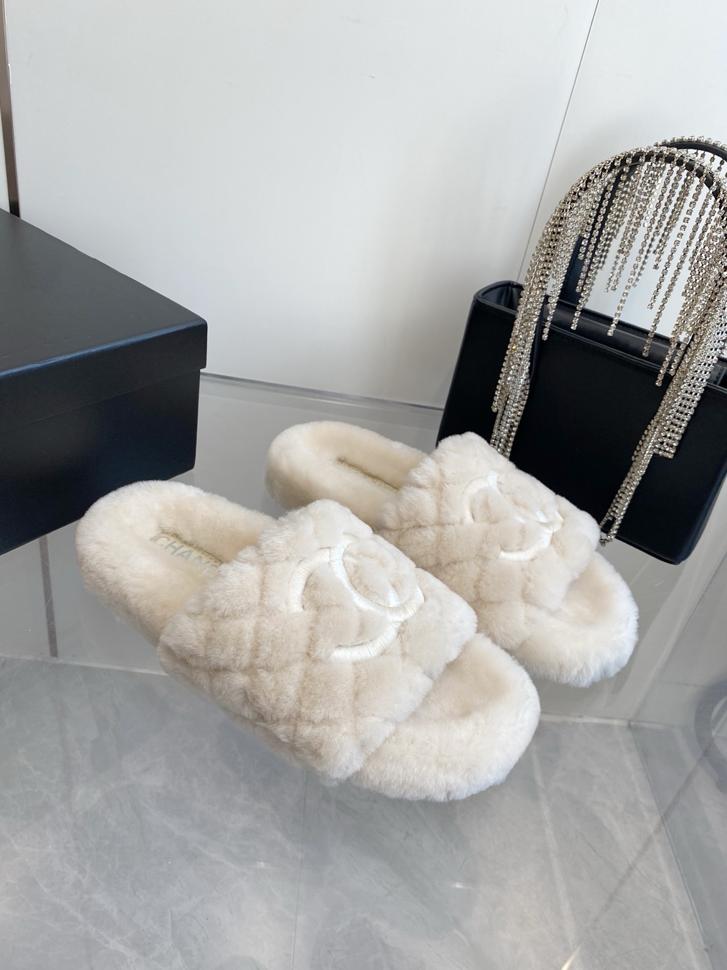 A pair of cream-colored luxury quilted faux fur slippers with an embroidered logo, placed next to a black handbag with crystal fringe. - Hình 2