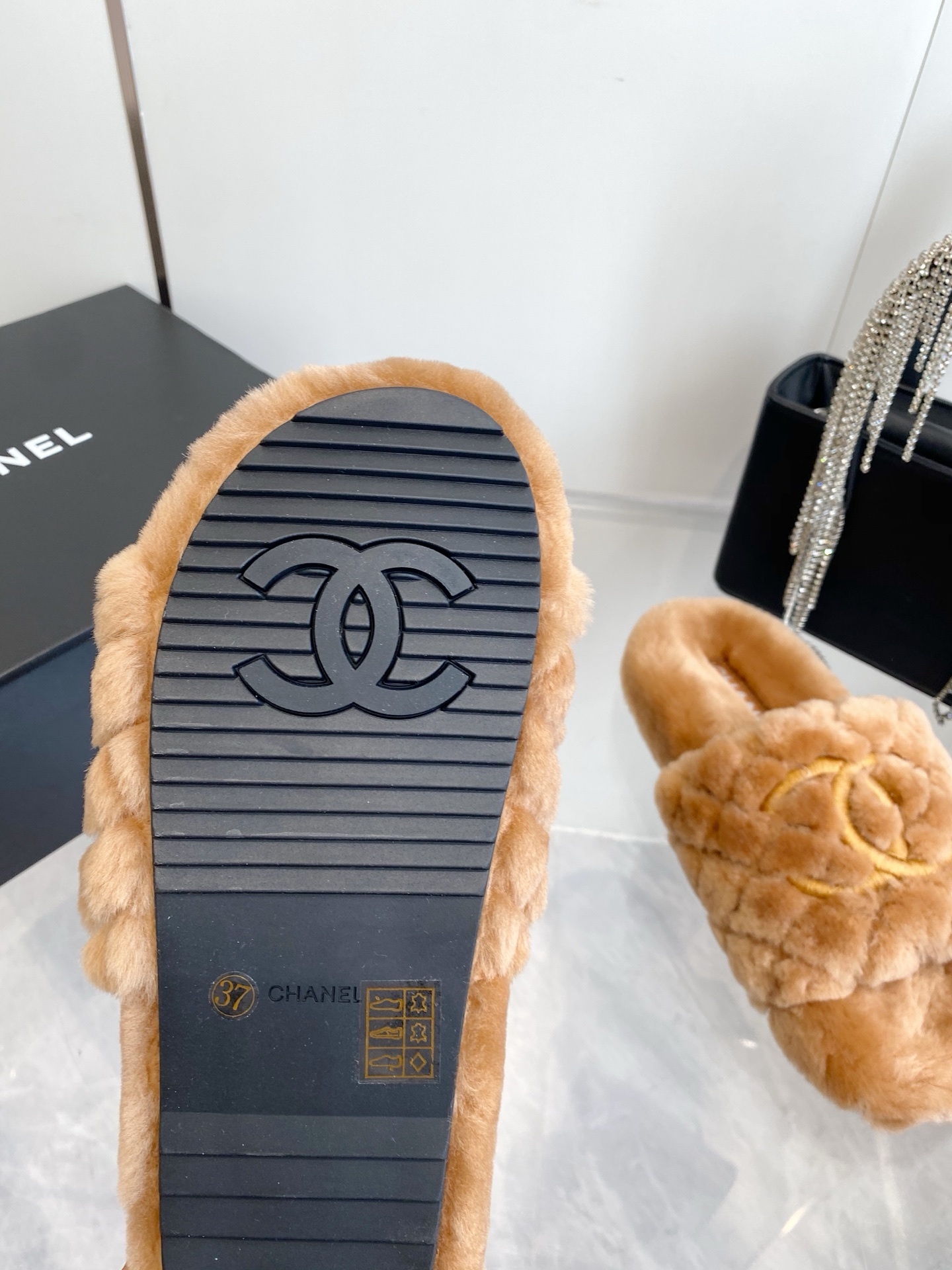A pair of camel-colored quilted shearling slides with embroidered interlocking CC logos on a marble surface next to a black handbag. - Hình 9