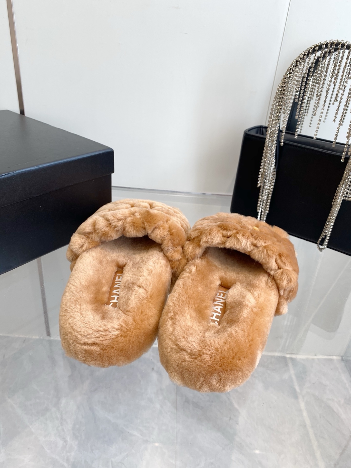 A pair of camel-colored quilted shearling slides with embroidered interlocking CC logos on a marble surface next to a black handbag. - Hình 8