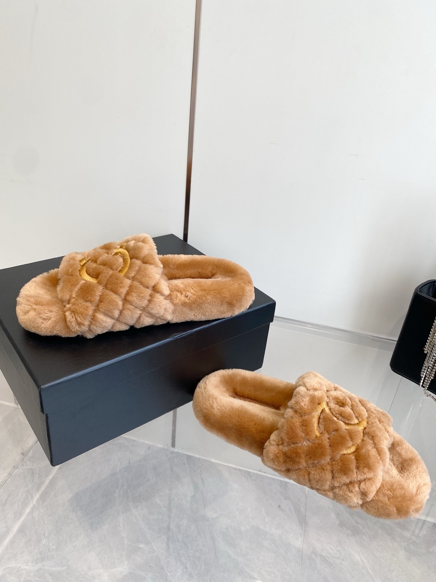 A pair of camel-colored quilted shearling slides with embroidered interlocking CC logos on a marble surface next to a black handbag. - Hình 7