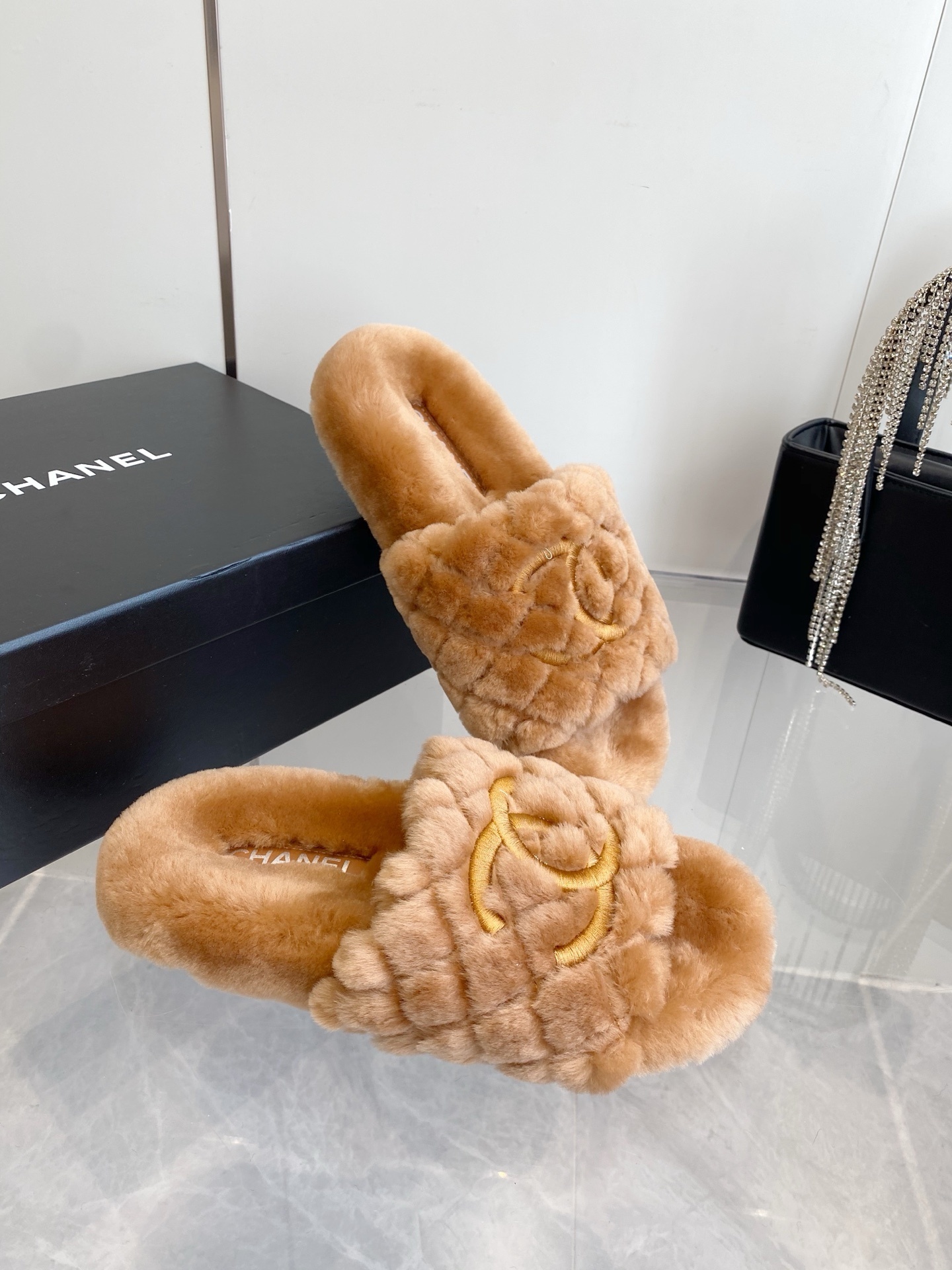 A pair of camel-colored quilted shearling slides with embroidered interlocking CC logos on a marble surface next to a black handbag. - Hình 6