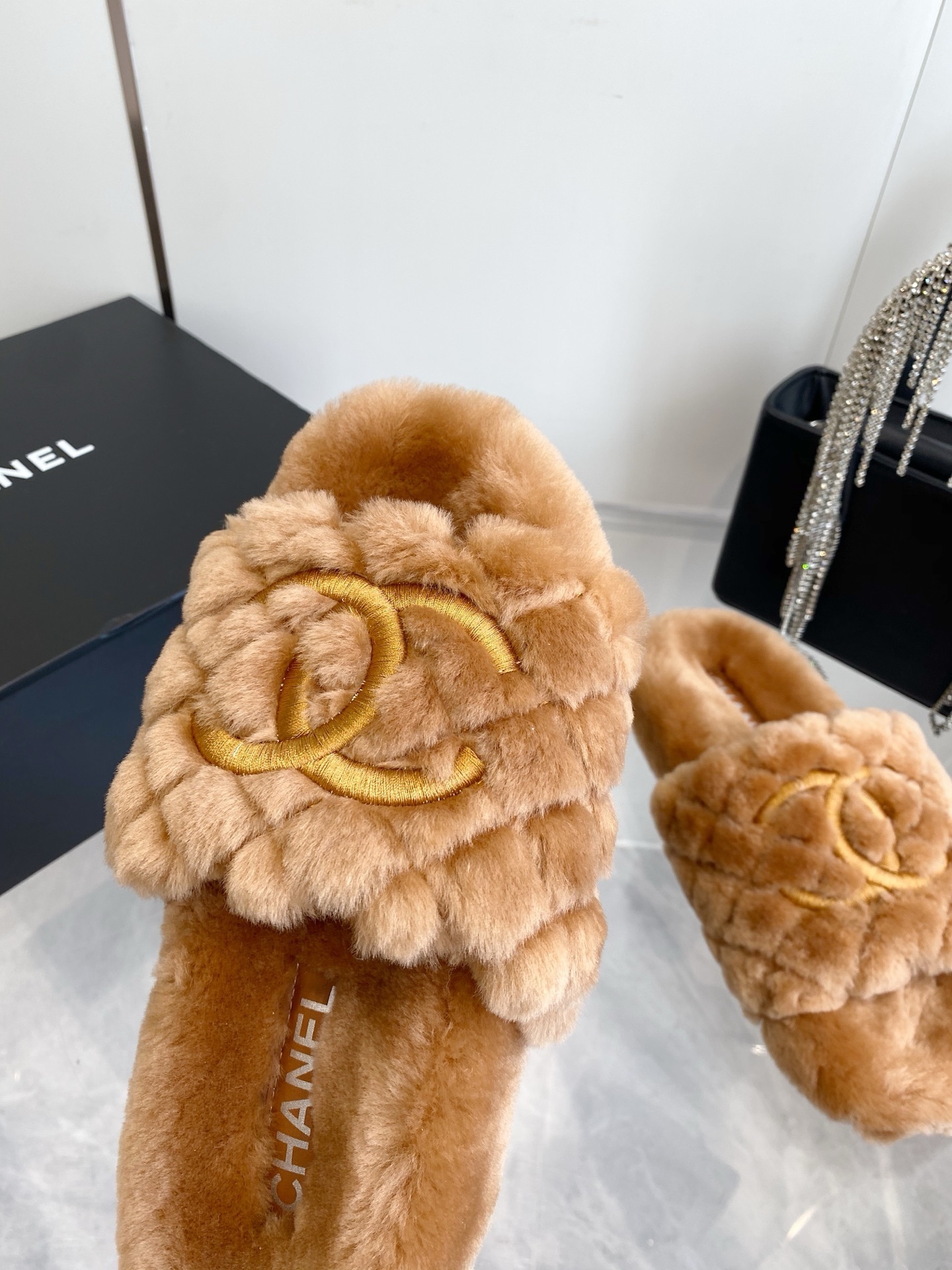 A pair of camel-colored quilted shearling slides with embroidered interlocking CC logos on a marble surface next to a black handbag. - Hình 5