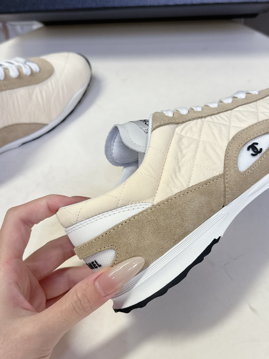 A pair of luxury Chanel quilted sneakers in beige and tan suede featuring the CC logo, placed next to a blue Chanel box. - Hình 8