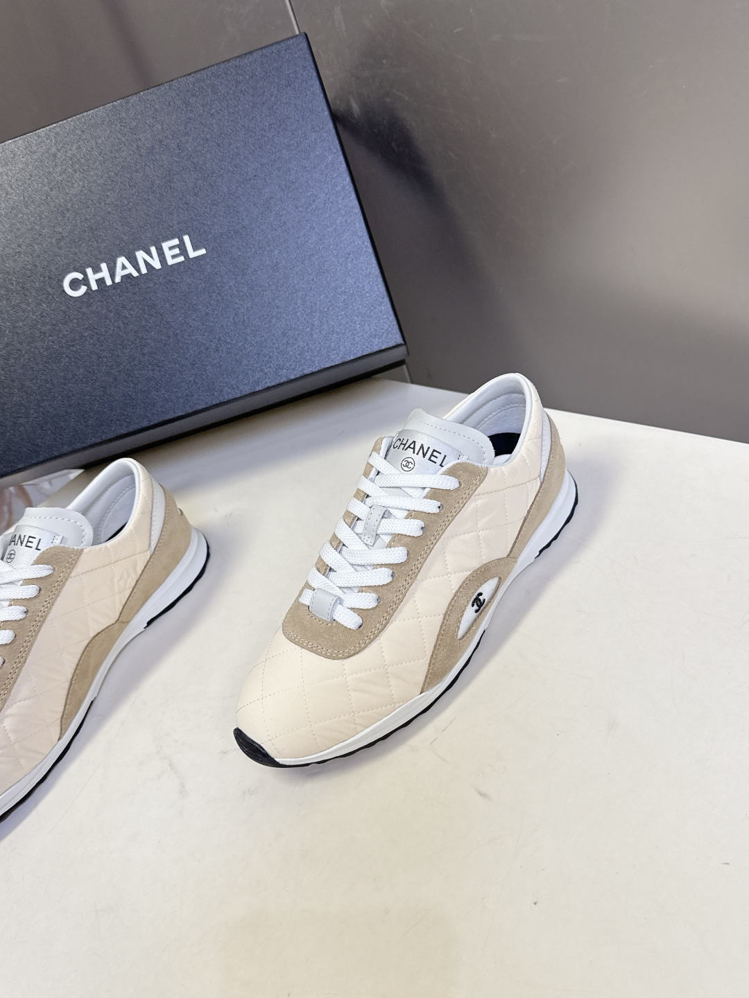 A pair of luxury Chanel quilted sneakers in beige and tan suede featuring the CC logo, placed next to a blue Chanel box. - Hình 3