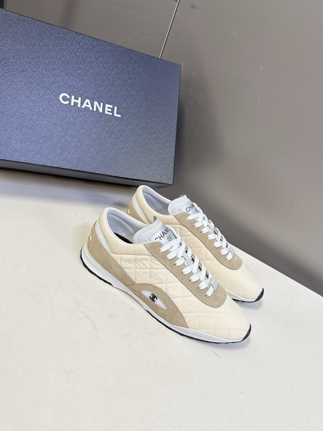 A pair of luxury Chanel quilted sneakers in beige and tan suede featuring the CC logo, placed next to a blue Chanel box. - Hình 2