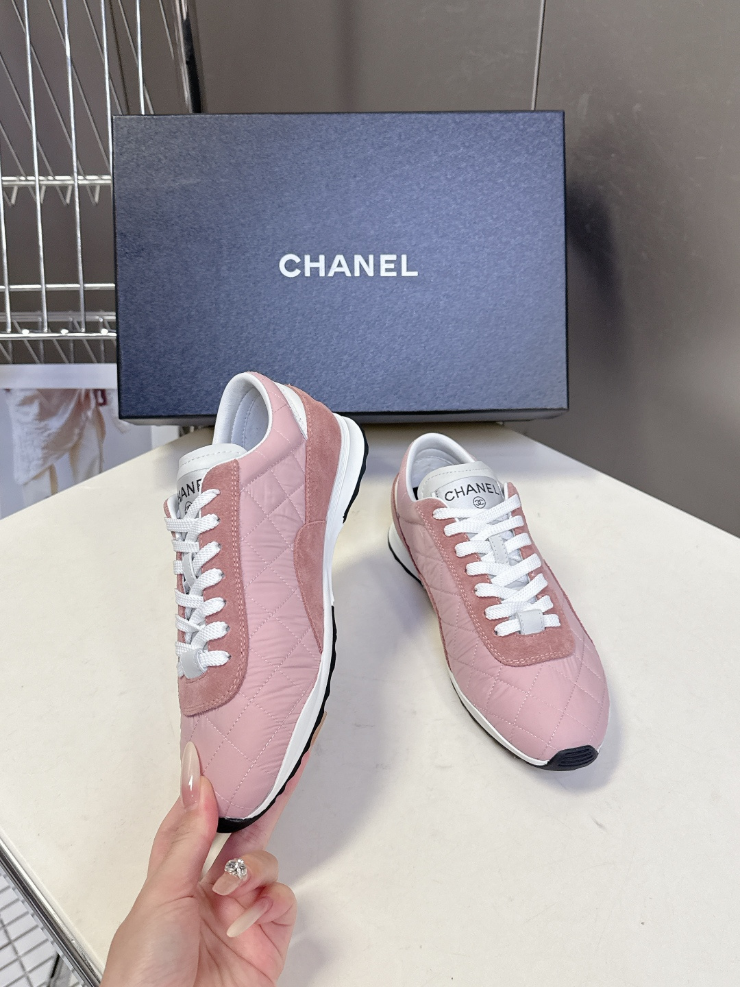 A pair of luxury pink quilted Chanel sneakers with white laces displayed next to their original black Chanel box. - Hình 4