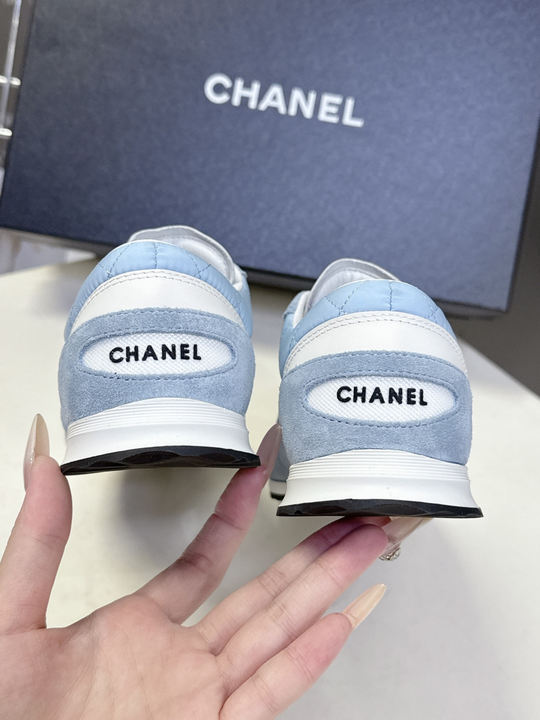 A pair of light blue Chanel quilted sneakers with white laces and suede accents next to a black Chanel shoe box. - Hình 9