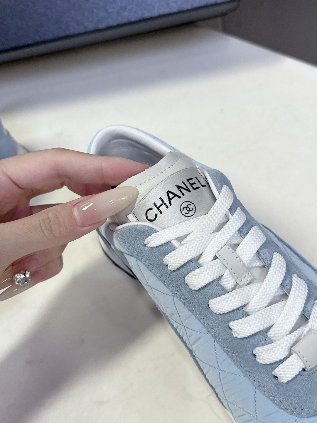 A pair of light blue Chanel quilted sneakers with white laces and suede accents next to a black Chanel shoe box. - Hình 7