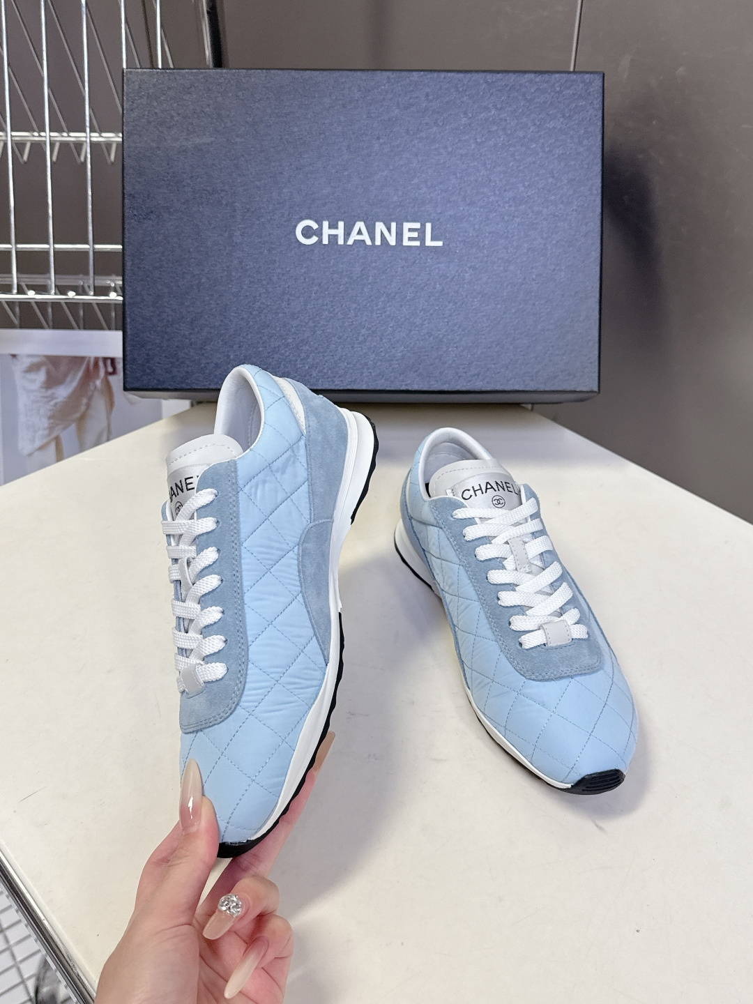 A pair of light blue Chanel quilted sneakers with white laces and suede accents next to a black Chanel shoe box. - Hình 4