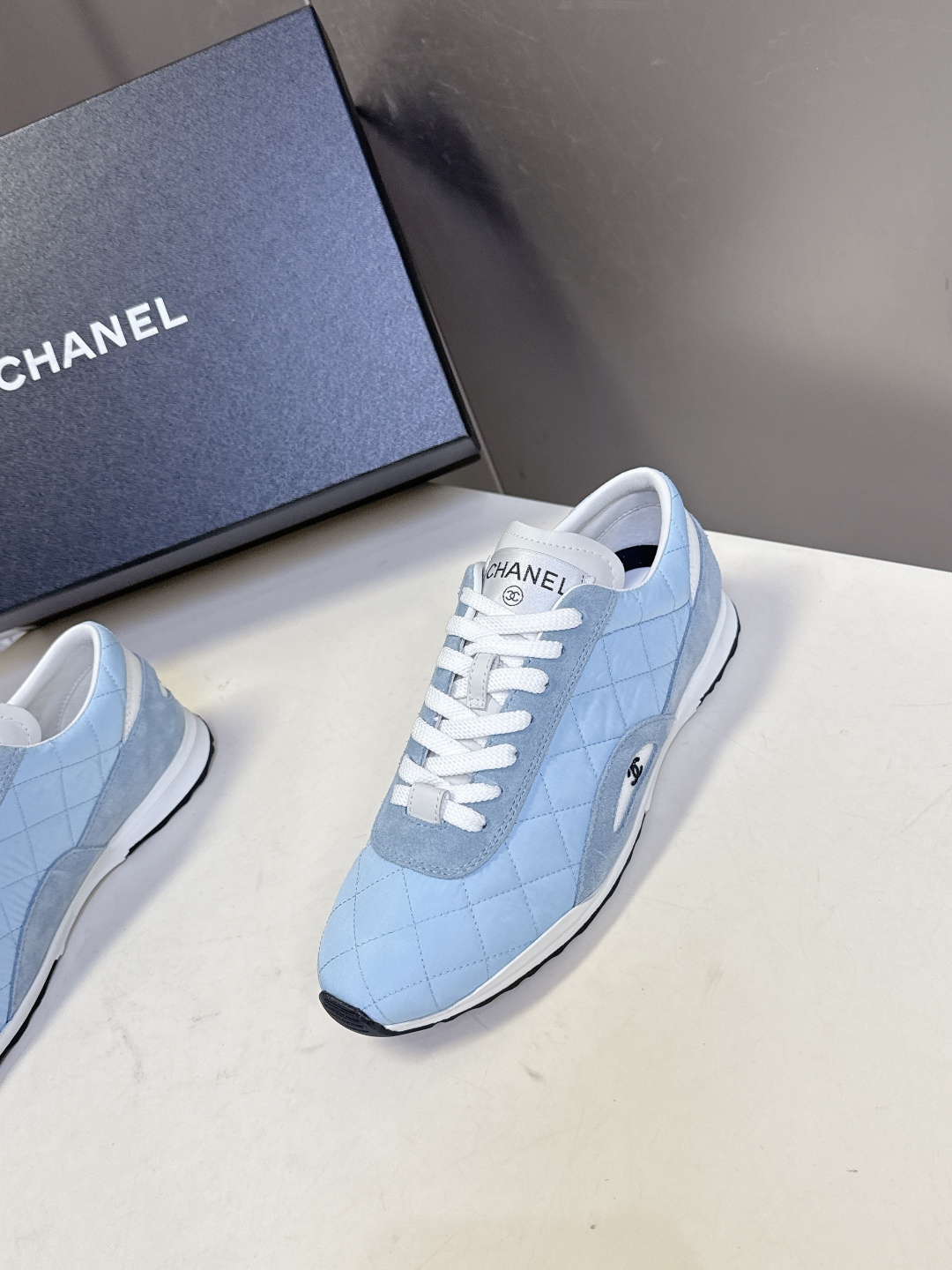 A pair of light blue Chanel quilted sneakers with white laces and suede accents next to a black Chanel shoe box. - Hình 3