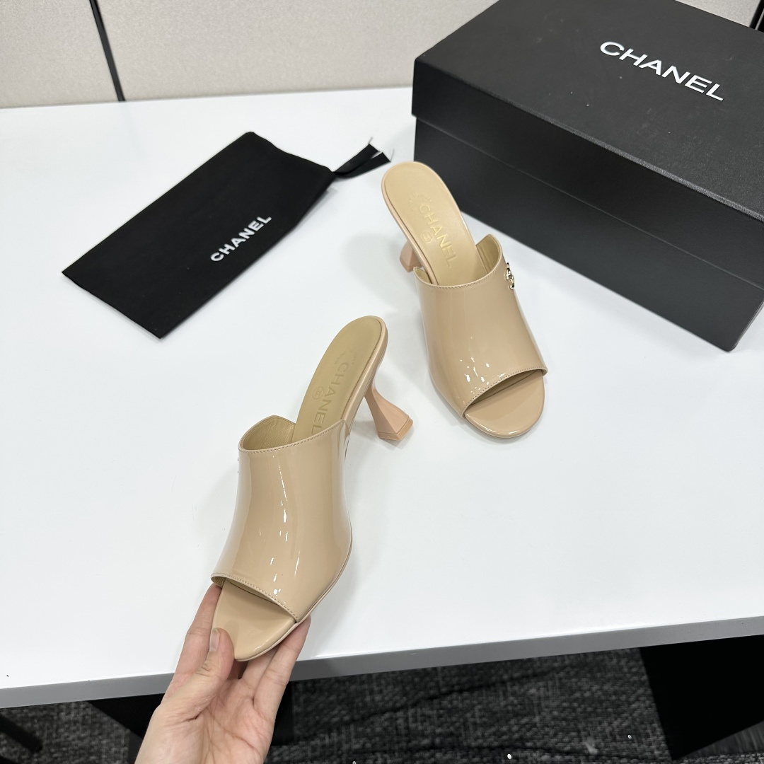 A pair of Chanel beige patent leather open-toe mules with gold CC logo and sculptural hourglass heels displayed on a black Chanel box. - Hình 2