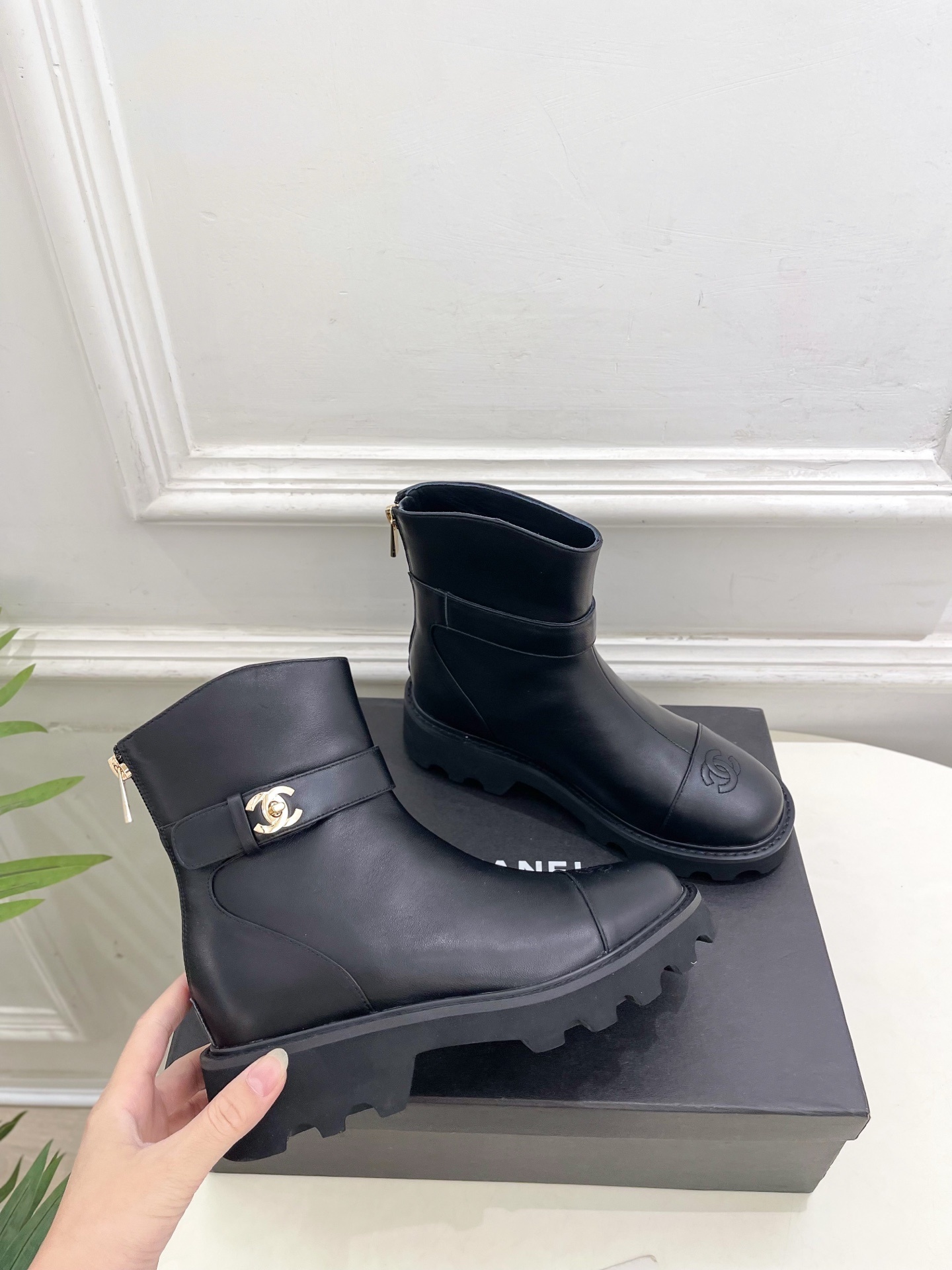 Pair of luxury black leather ankle boots with gold CC logo buckle and chunky lug sole displayed on a black box. - Hình 4