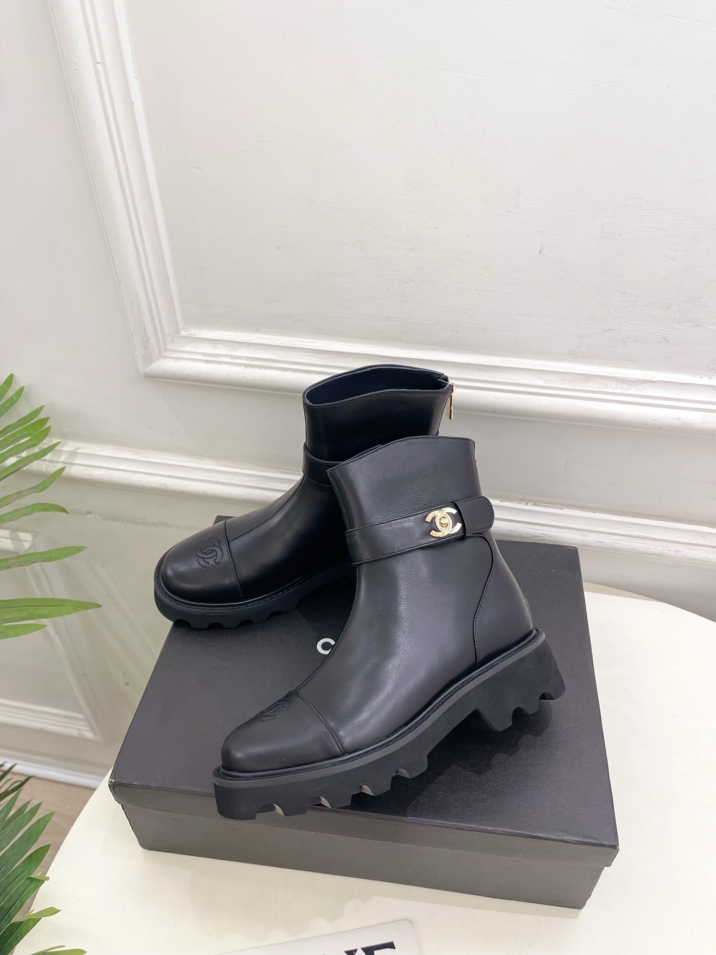 Pair of luxury black leather ankle boots with gold CC logo buckle and chunky lug sole displayed on a black box. - Hình 3