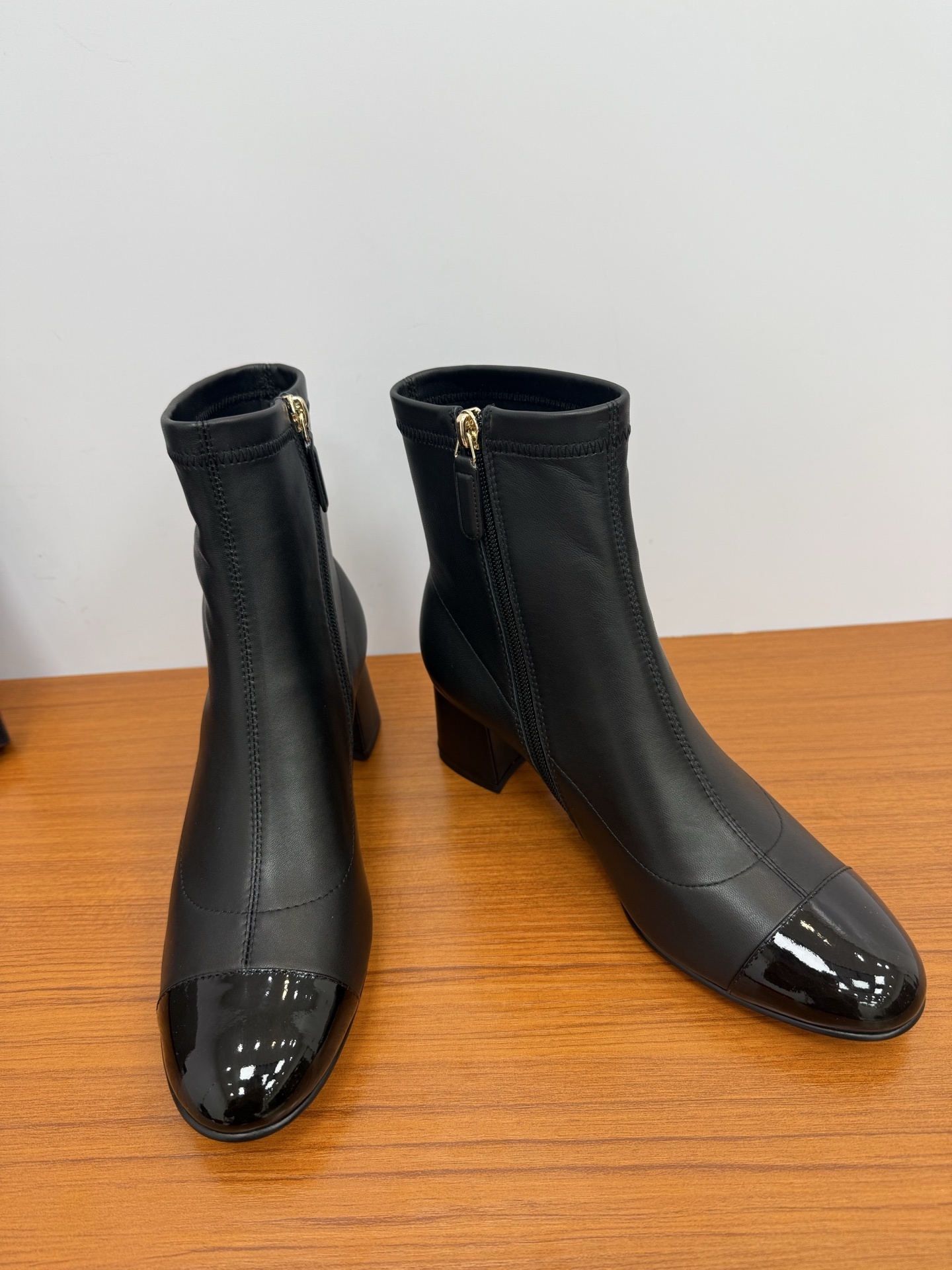 Pair of elegant black leather ankle boots with glossy patent toe caps and gold-tone zipper hardware on a wooden surface. - Hình 7