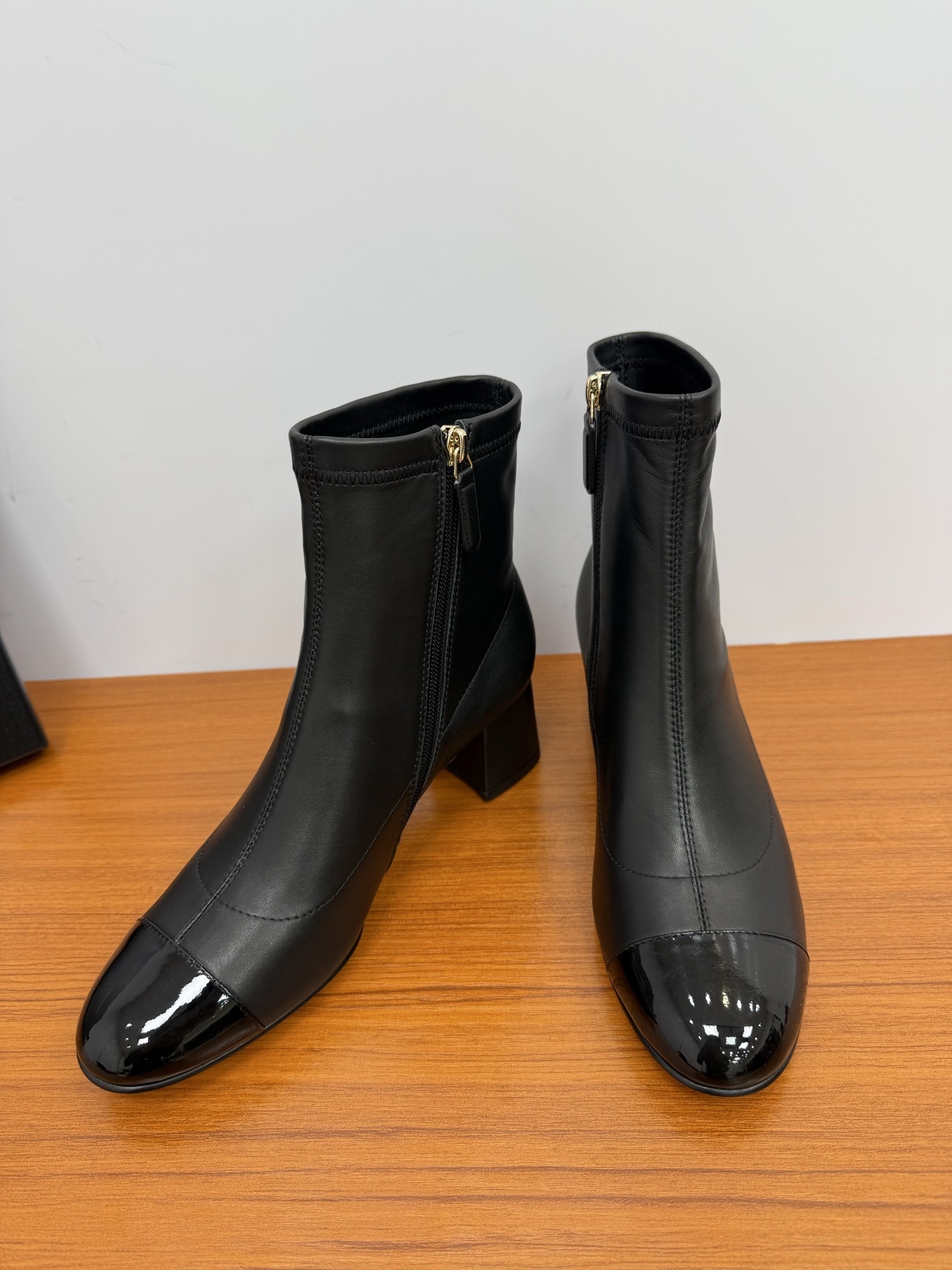 Pair of elegant black leather ankle boots with glossy patent toe caps and gold-tone zipper hardware on a wooden surface. - Hình 6