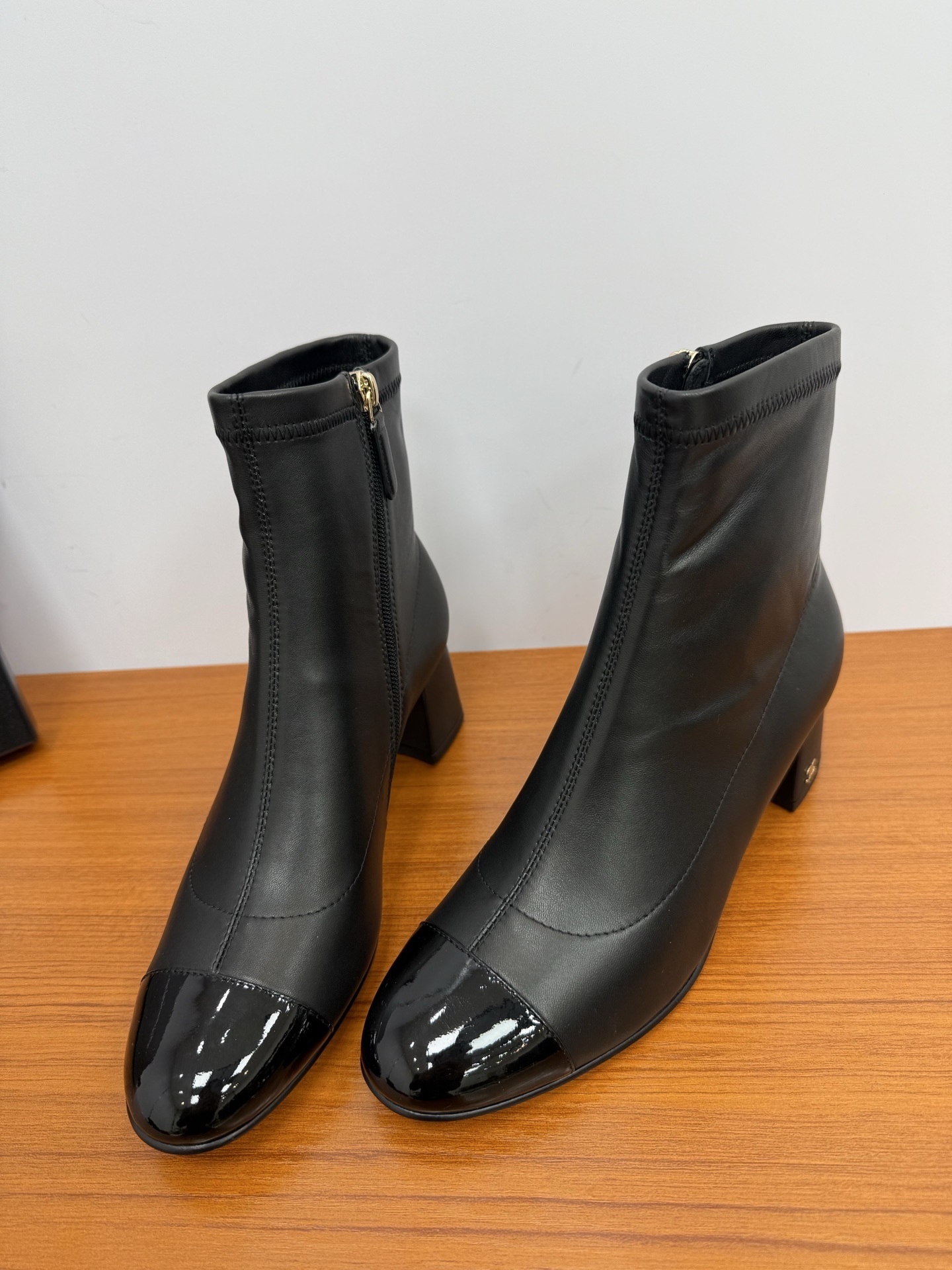 Pair of elegant black leather ankle boots with glossy patent toe caps and gold-tone zipper hardware on a wooden surface. - Hình 5