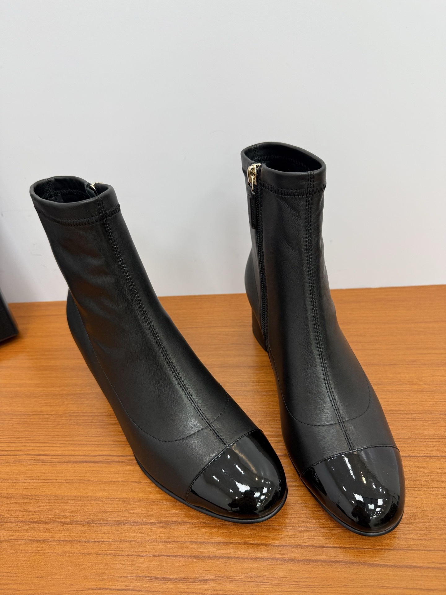 Pair of elegant black leather ankle boots with glossy patent toe caps and gold-tone zipper hardware on a wooden surface. - Hình 4