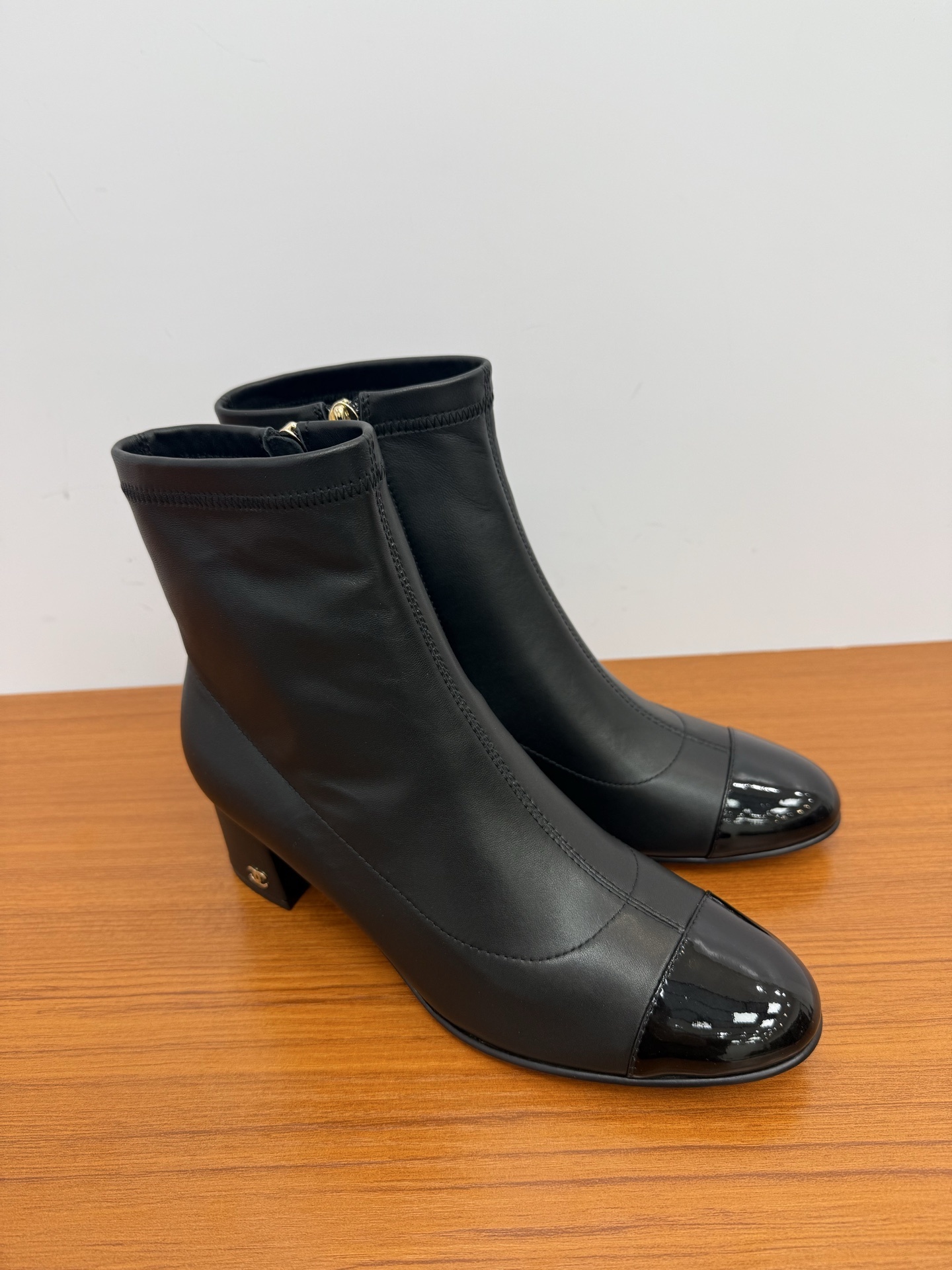 Pair of elegant black leather ankle boots with glossy patent toe caps and gold-tone zipper hardware on a wooden surface. - Hình 2