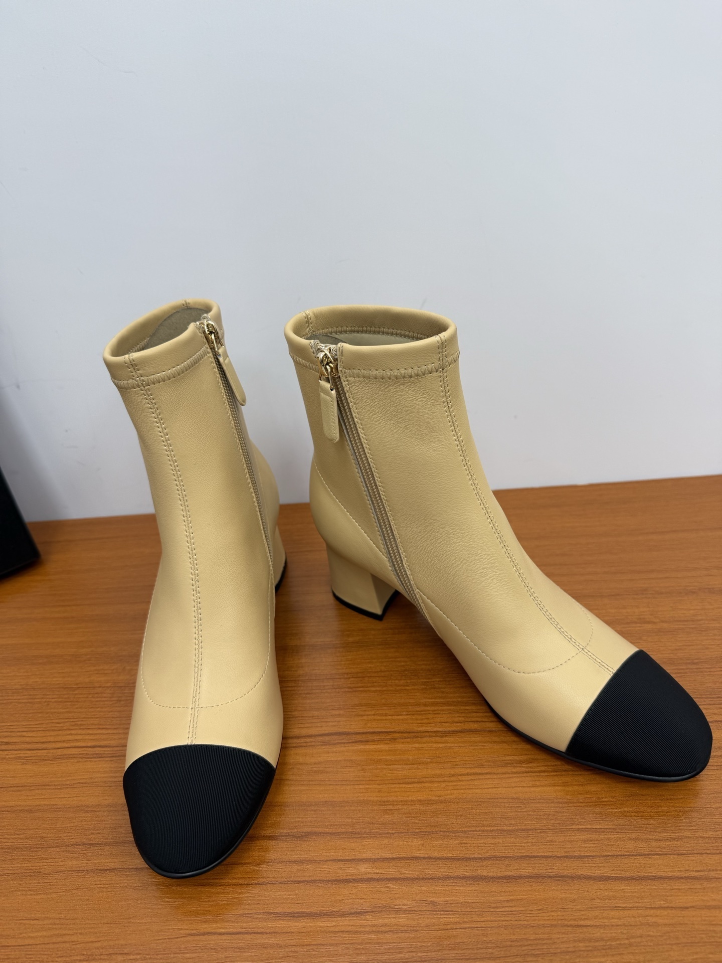 Pair of beige leather ankle boots with black fabric cap toes and side zippers on a wooden surface. - Hình 6
