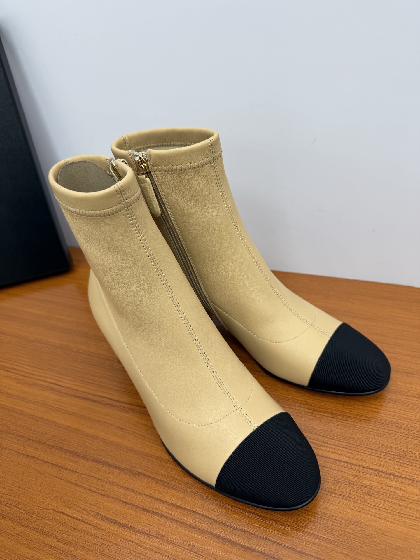 Pair of beige leather ankle boots with black fabric cap toes and side zippers on a wooden surface. - Hình 2