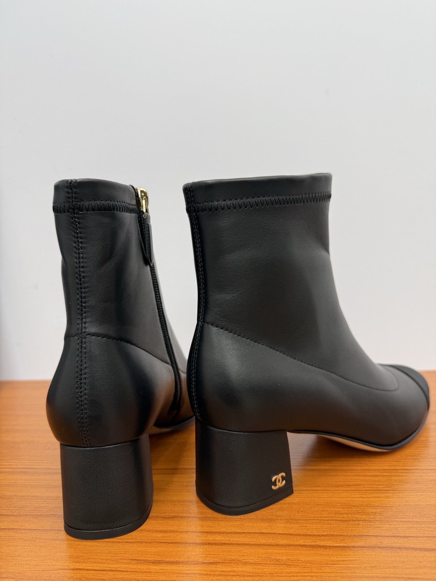 Pair of black leather ankle boots with a fabric cap toe and gold-tone side zipper on a wooden surface. - Hình 8