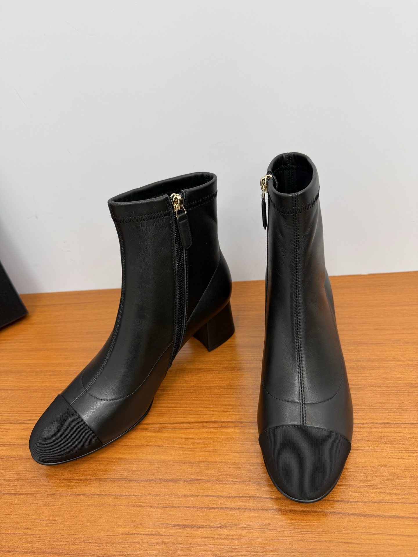 Pair of black leather ankle boots with a fabric cap toe and gold-tone side zipper on a wooden surface. - Hình 6