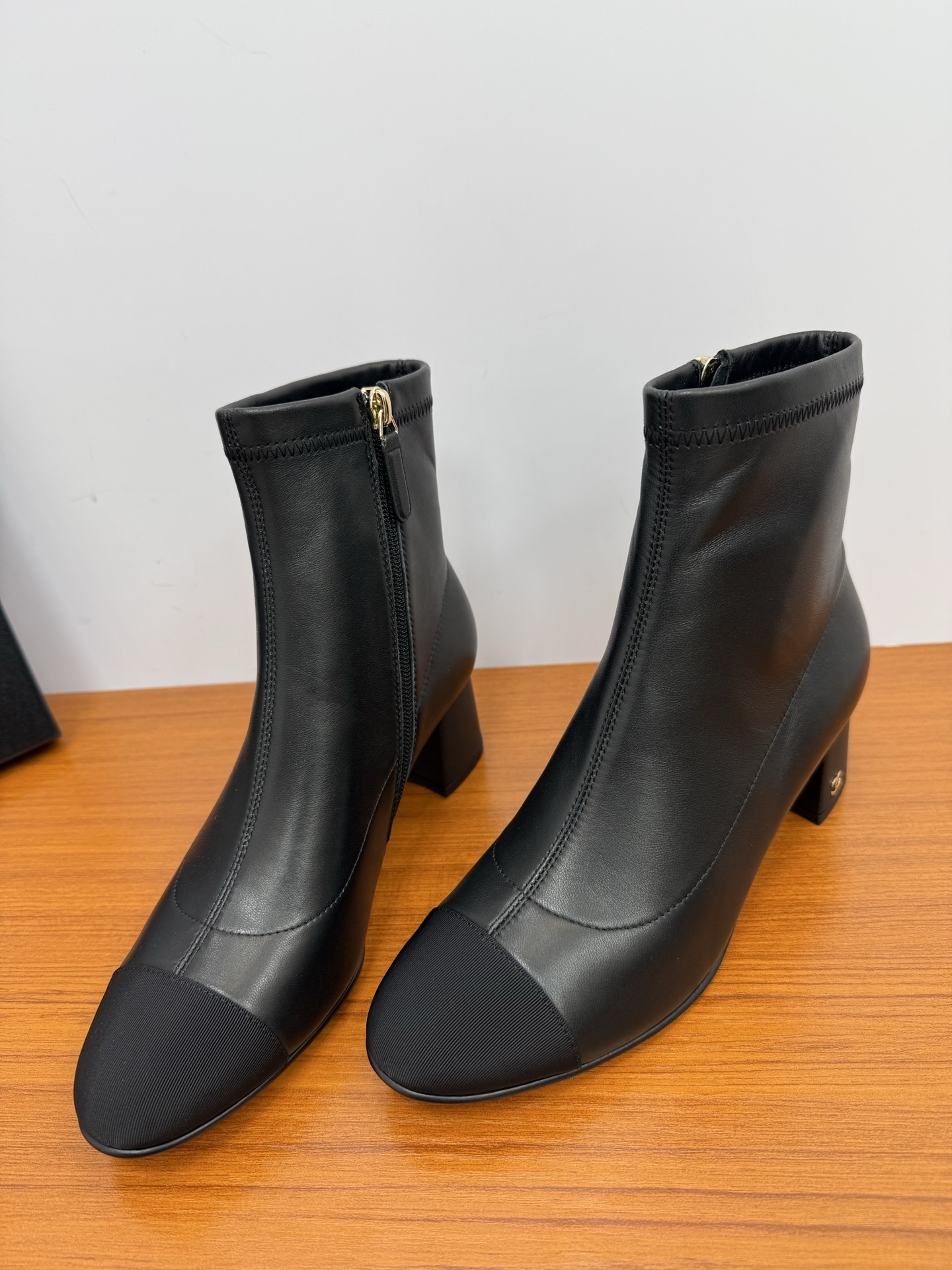 Pair of black leather ankle boots with a fabric cap toe and gold-tone side zipper on a wooden surface. - Hình 5