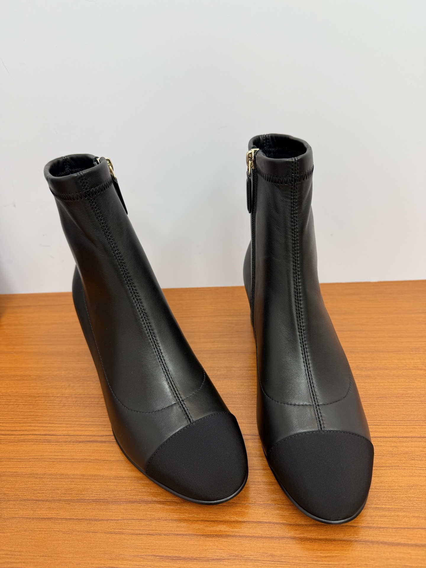 Pair of black leather ankle boots with a fabric cap toe and gold-tone side zipper on a wooden surface. - Hình 4