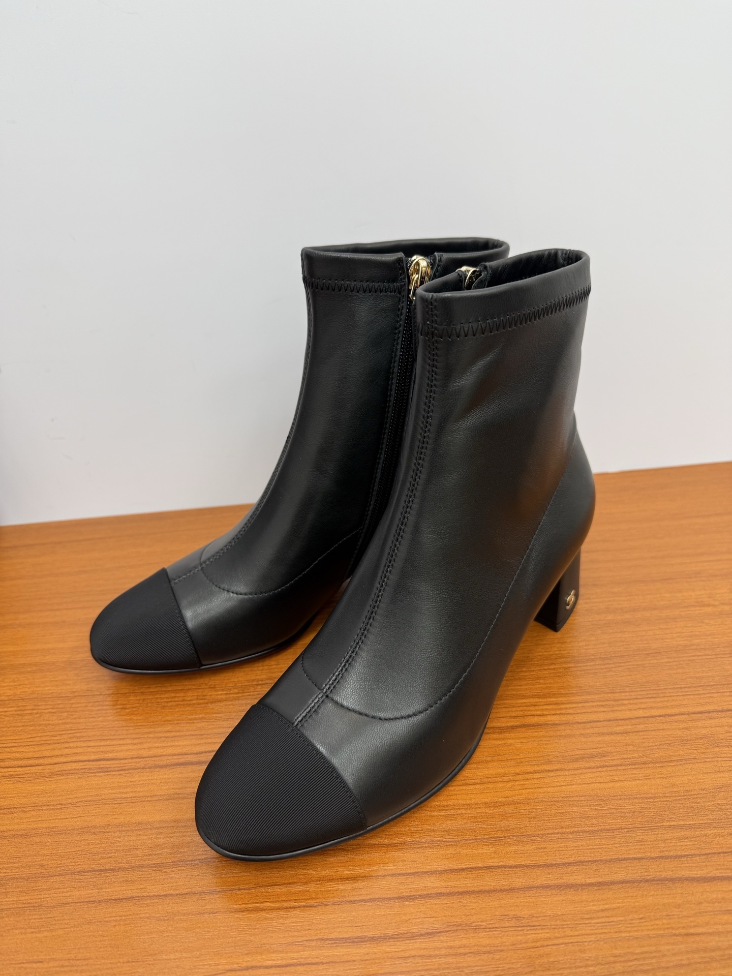 Pair of black leather ankle boots with a fabric cap toe and gold-tone side zipper on a wooden surface. - Hình 3