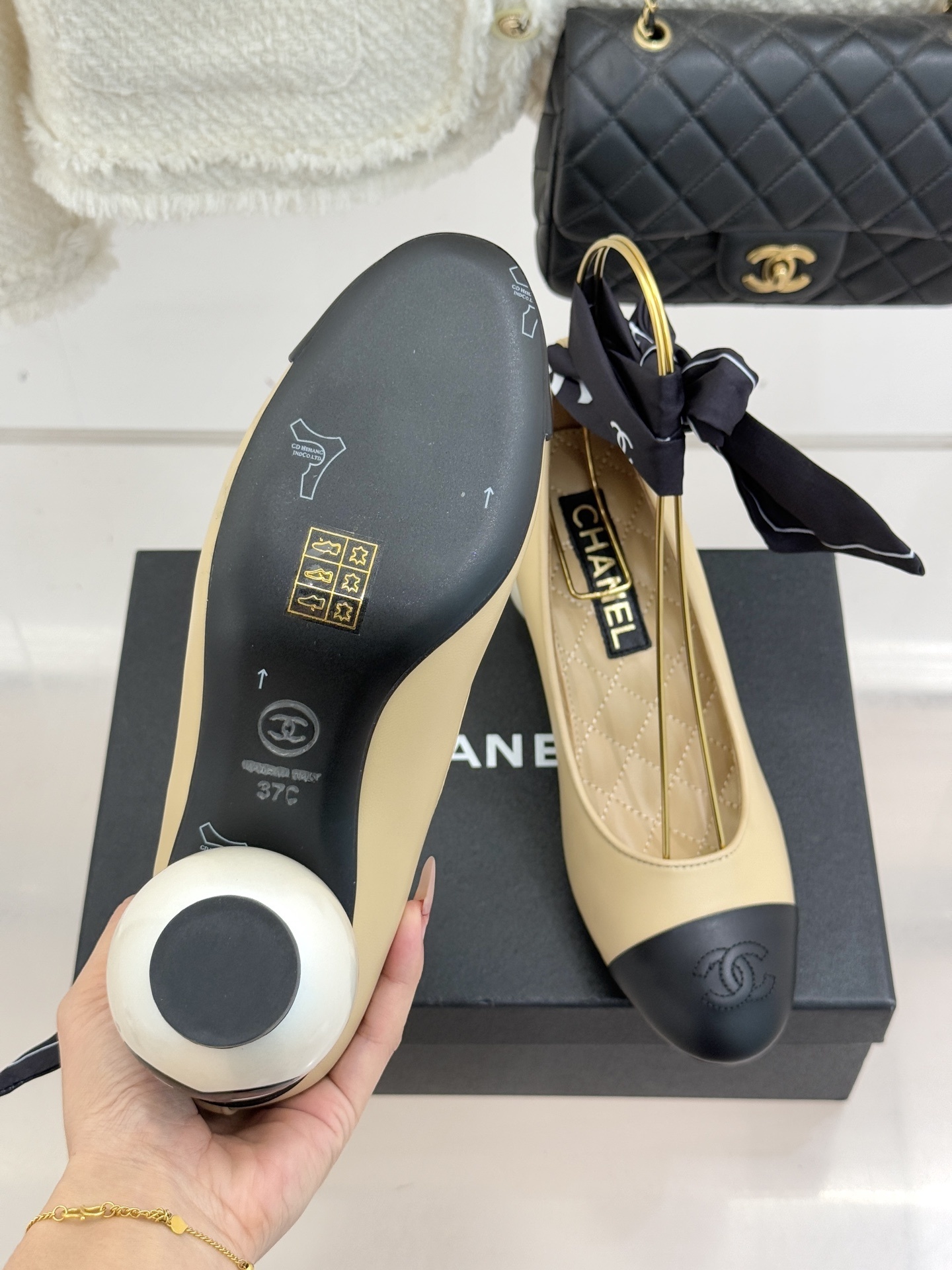 Pair of Chanel beige leather pumps with black cap toes, unique pearl-shaped heels, and black silk ribbons displayed on a black box. - Hình 9