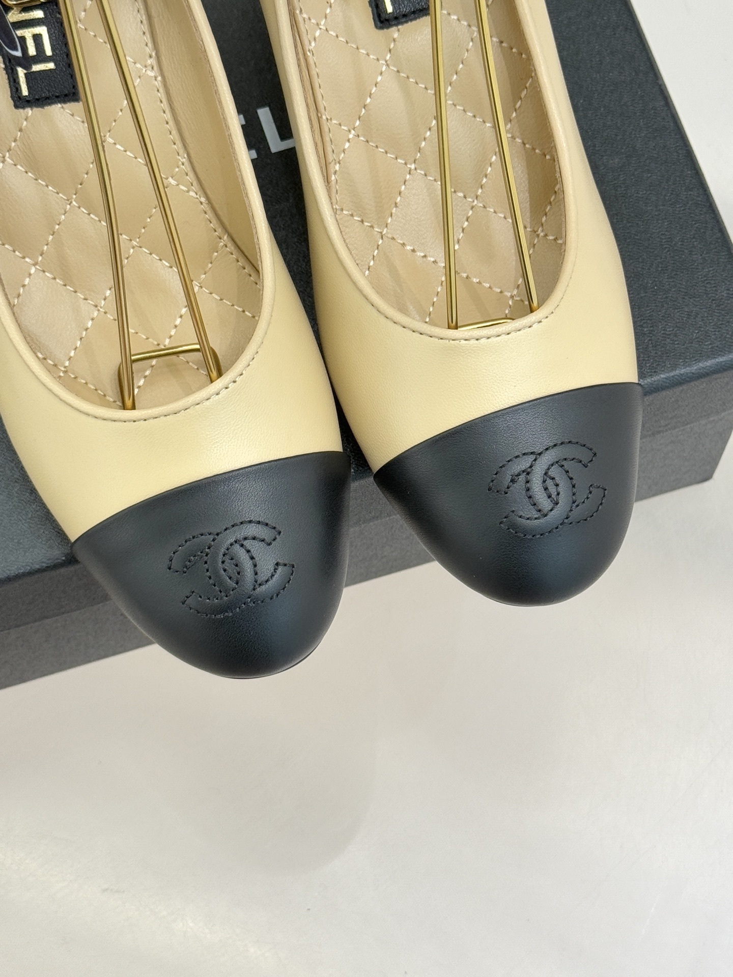 Pair of Chanel beige leather pumps with black cap toes, unique pearl-shaped heels, and black silk ribbons displayed on a black box. - Hình 7