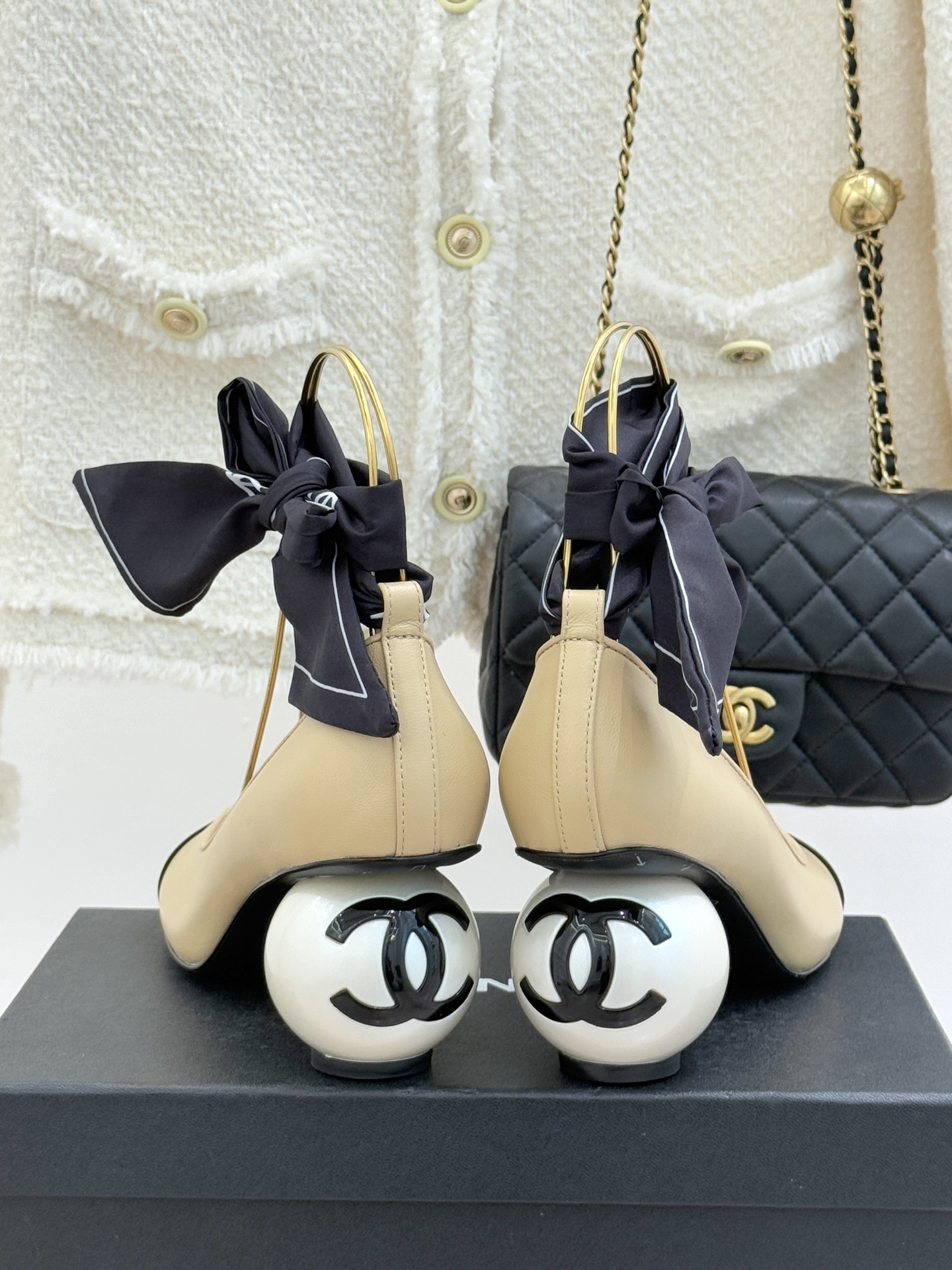 Pair of Chanel beige leather pumps with black cap toes, unique pearl-shaped heels, and black silk ribbons displayed on a black box. - Hình 6