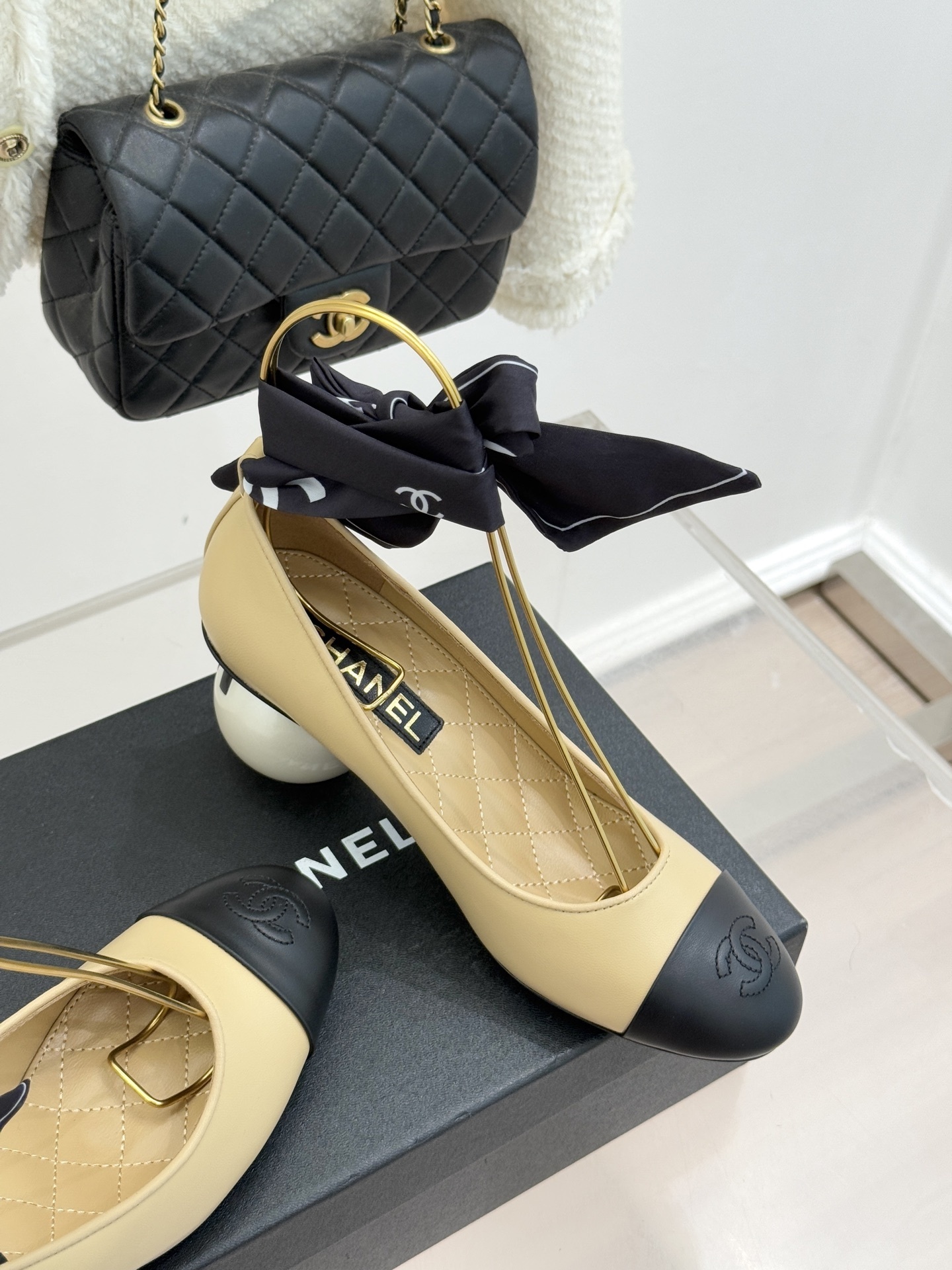 Pair of Chanel beige leather pumps with black cap toes, unique pearl-shaped heels, and black silk ribbons displayed on a black box. - Hình 5