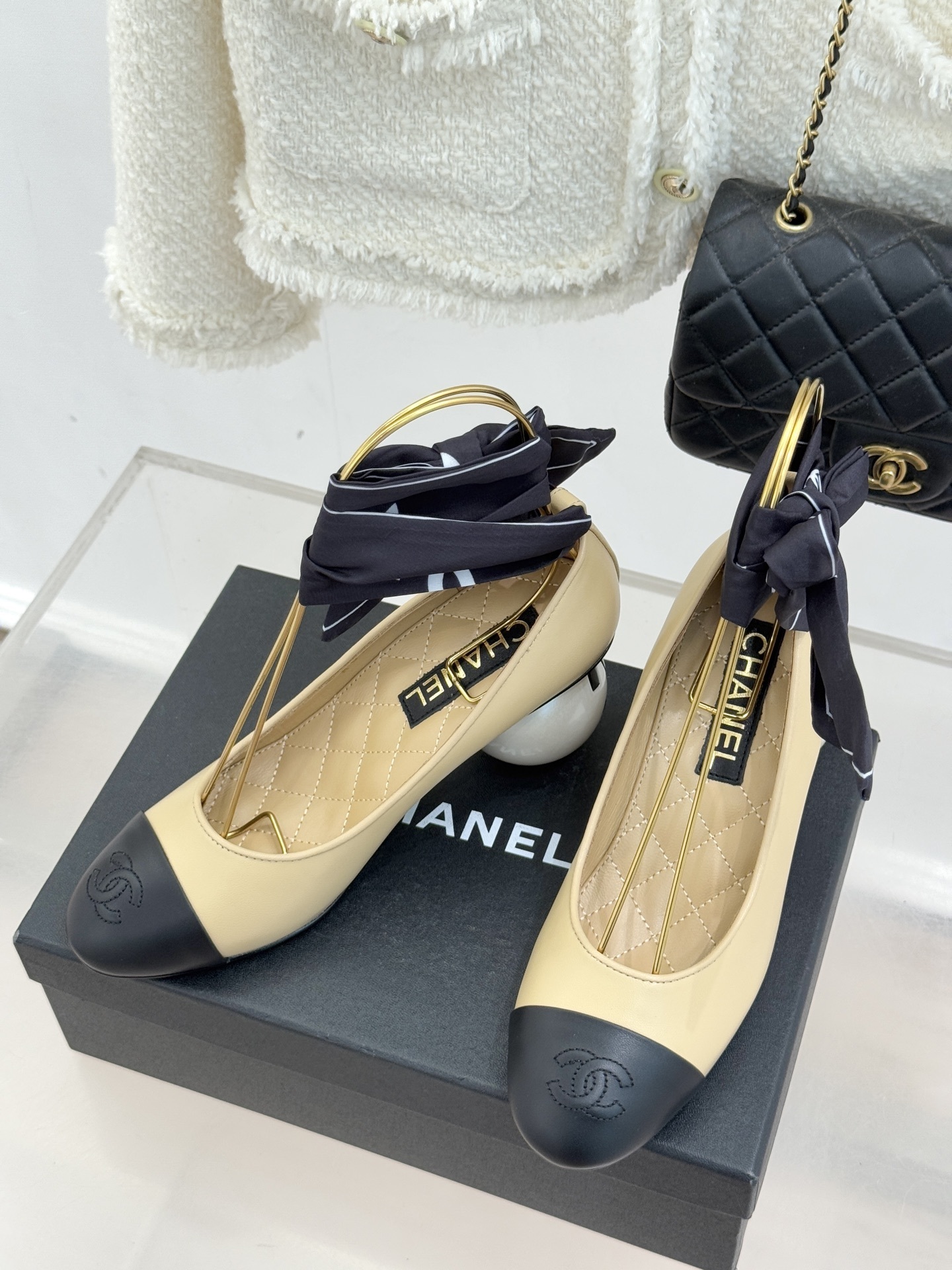 Pair of Chanel beige leather pumps with black cap toes, unique pearl-shaped heels, and black silk ribbons displayed on a black box. - Hình 2
