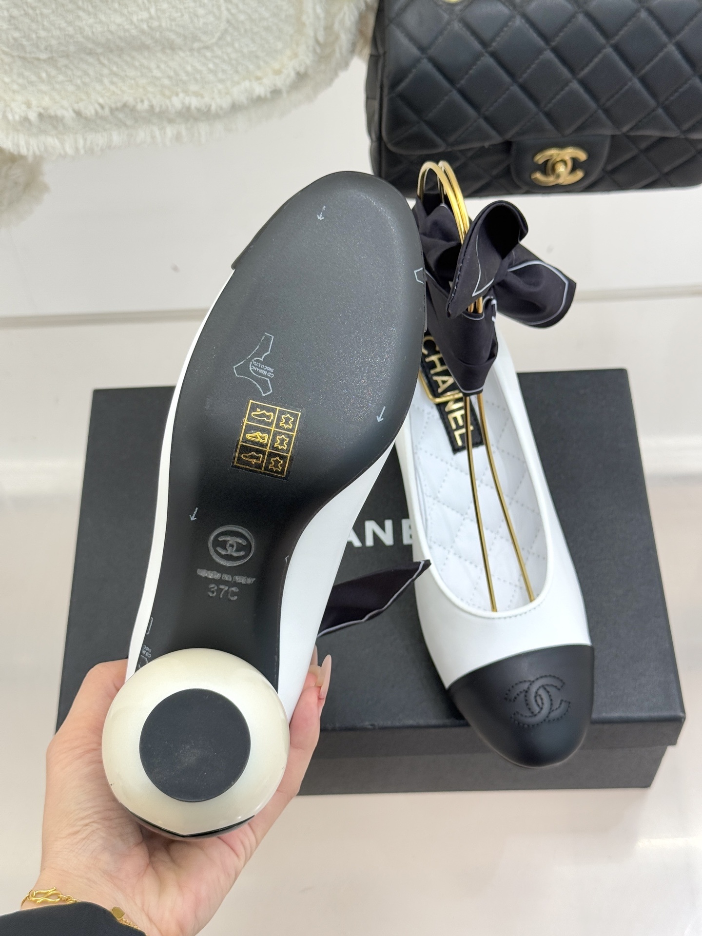 Elegant Chanel two-tone white and black pumps with a round pearl heel and black silk ribbon ankle bows displayed on a black box. - Hình 9