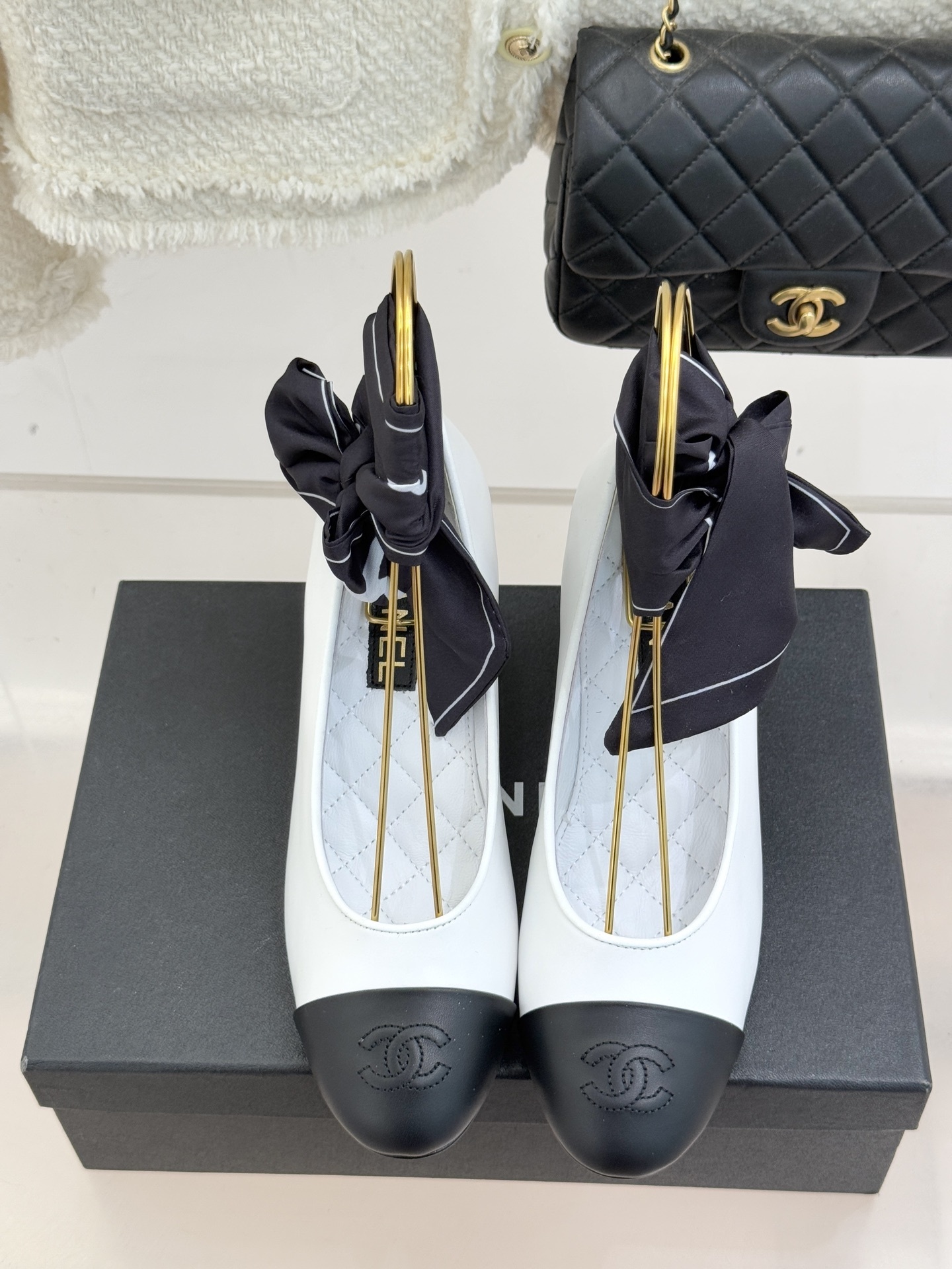 Elegant Chanel two-tone white and black pumps with a round pearl heel and black silk ribbon ankle bows displayed on a black box. - Hình 4