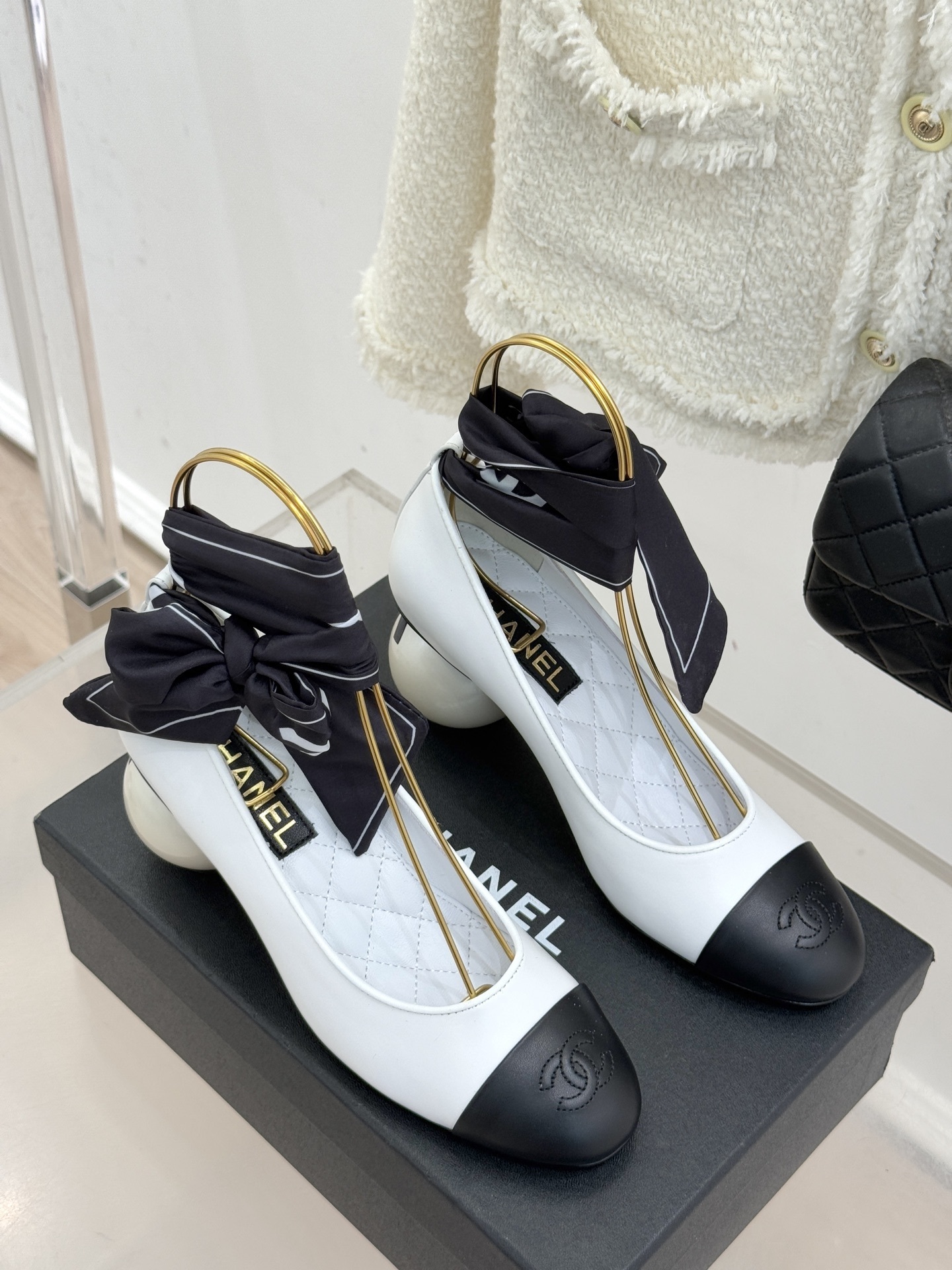 Elegant Chanel two-tone white and black pumps with a round pearl heel and black silk ribbon ankle bows displayed on a black box. - Hình 3
