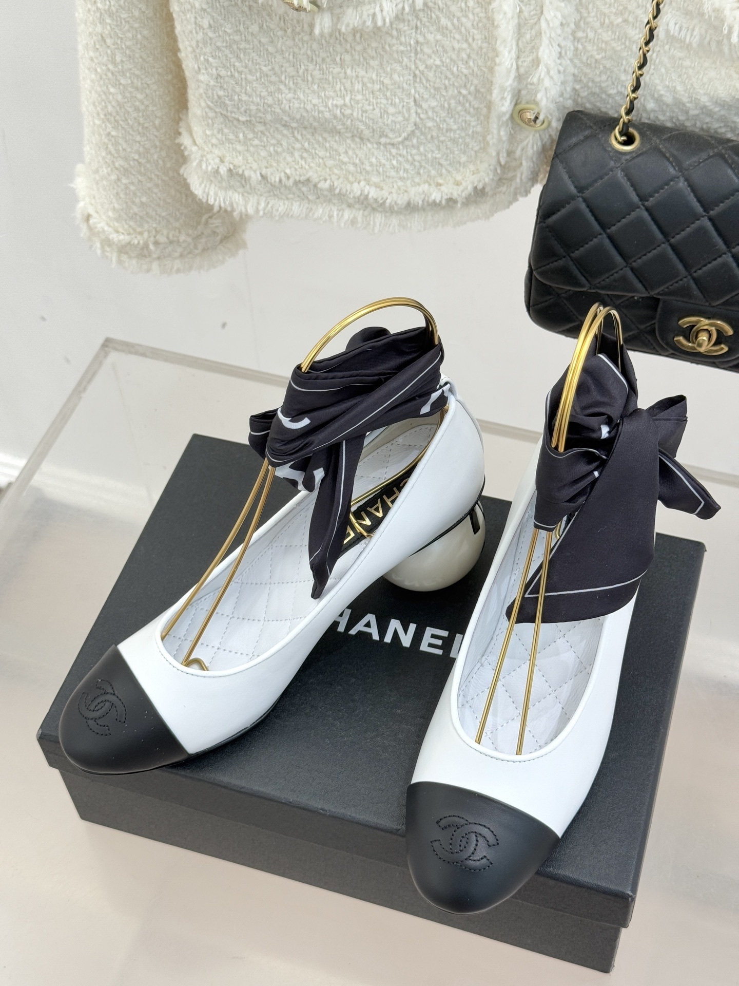 Elegant Chanel two-tone white and black pumps with a round pearl heel and black silk ribbon ankle bows displayed on a black box. - Hình 2